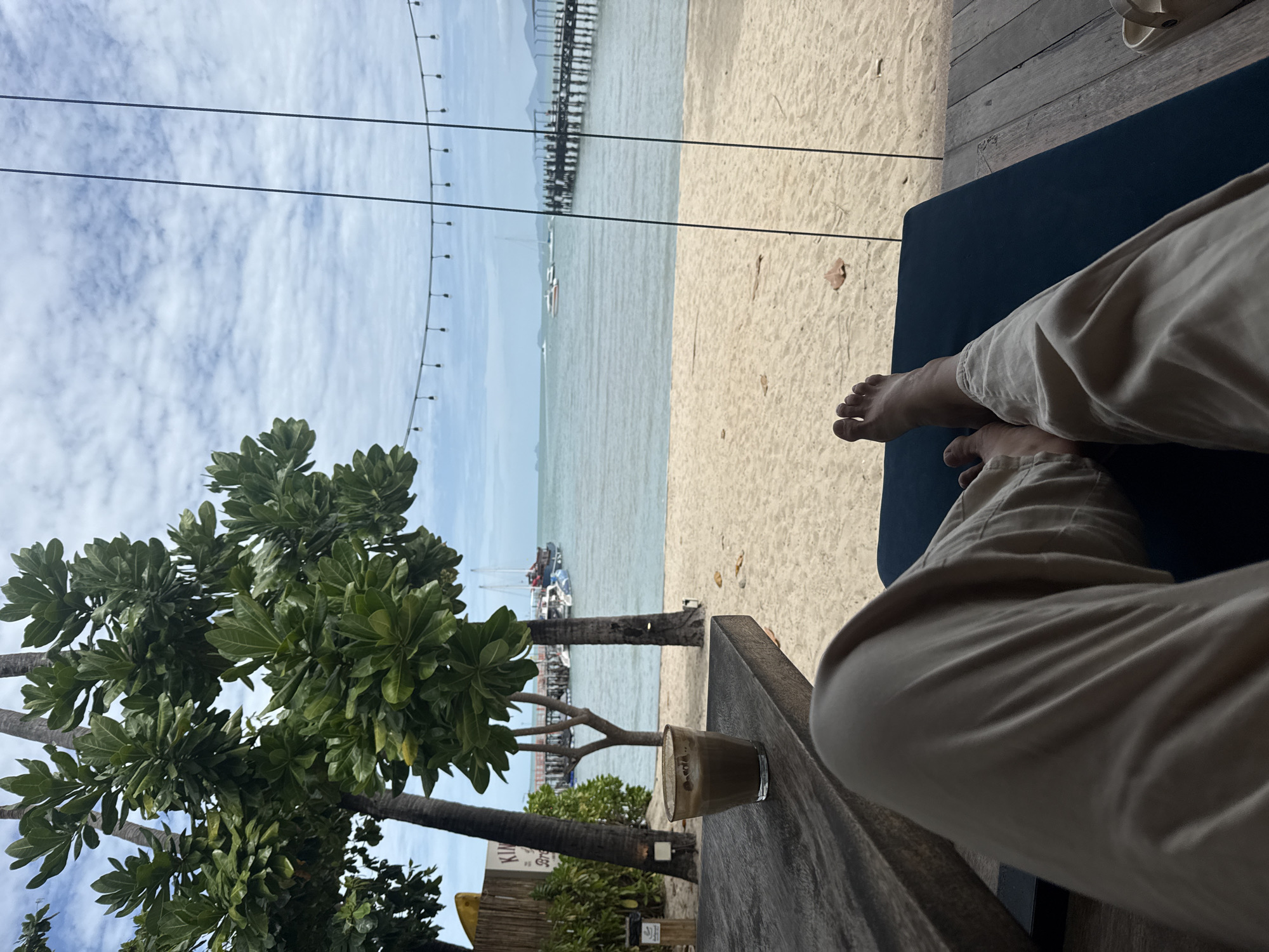 The image captures a serene beachside scene in the Ko Samui District of Thailand. In the foreground, a person's legs, clad in light-colored pants, are visible as they recline on a dark blue cushioned lounger. Their bare feet are at the edge of the lounger, with one foot slightly lifted. To the left, a rustic wooden table holds a glass of coffee, its surface showing a frothy texture. Beyond the table and lounger, a sandy beach stretches towards the calm, blue ocean. Palm trees with lush green foliage are interspersed with slender tree trunks, casting shadows on the sand. In the background, a wooden pier extends into the water, with several boats anchored nearby. The sky is partially cloudy, with a string of decorative lights strung across the upper frame, adding a touch of ambiance. The overall atmosphere is one of relaxation and enjoyment of a tropical paradise.