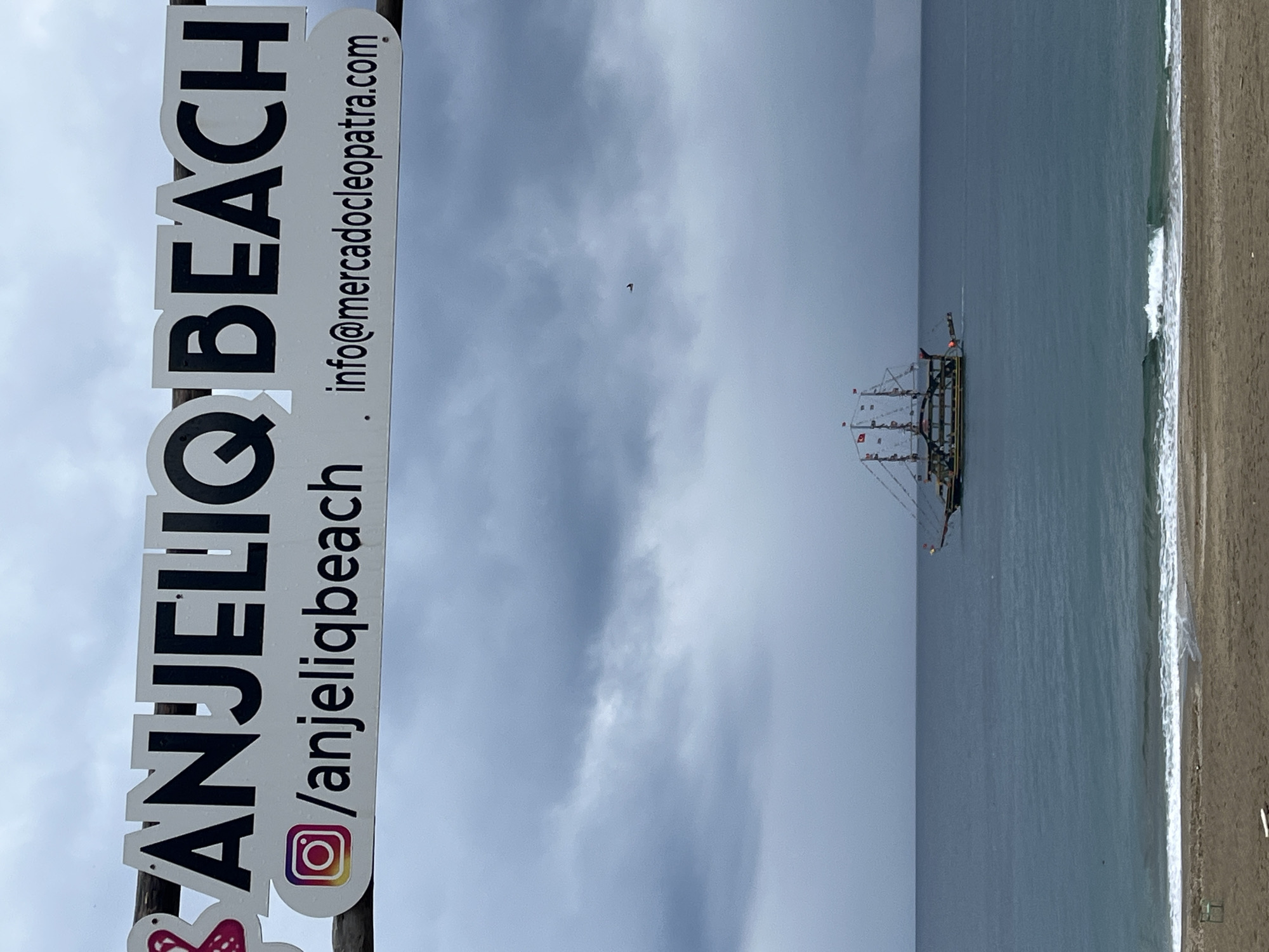 The image depicts a coastal scene with a sign in the foreground that reads "Anjeliq Beach" vertically in large letters, accompanied by an Instagram handle "/anjeliqbeach" and an email address "info@mercadocleopatra.com." Beyond the sign, there's a seascape featuring a sailing ship positioned off the coast. The shoreline is visible with waves meeting the sandy beach. The sky is overcast, which suggests either dawn or dusk. The location is indicated by the sign as "Anjeliq Beach," known to be situated in the city of Alanya, Turkey.