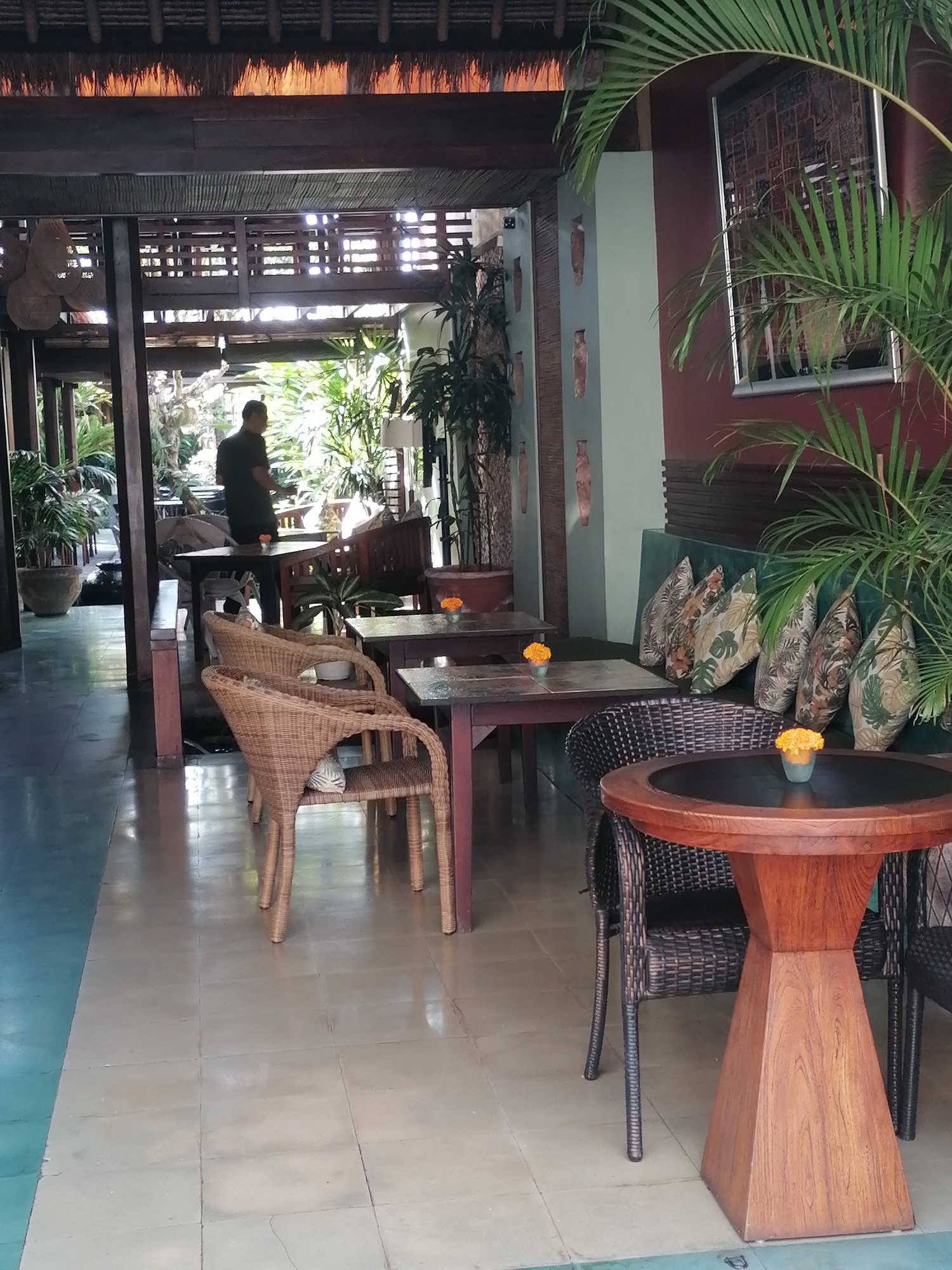 The image depicts the interior of an open-air cafe or restaurant in Ubud, Indonesia. The setting features a rustic, tropical ambiance with natural materials. Woven rattan chairs are arranged around wooden tables. In the foreground, a round wooden table with a distinctive geometric base stands. Behind it, a cushioned bench is adorned with floral-patterned pillows.
A person, appearing to be a male with short hair, is visible in the mid-ground, standing near a table. He is wearing a dark, possibly green, polo shirt and dark trousers. The floor is tiled, with a section transitioning to a different color near the left edge of the frame. Lush greenery, including palm fronds, frames the scene, suggesting an outdoor or semi-outdoor space. The architecture incorporates wooden beams and thatched roofing elements, common in Balinese design. Vases are displayed on a wall in the background. The lighting appears to be natural, coming from the open sides of the establishment.