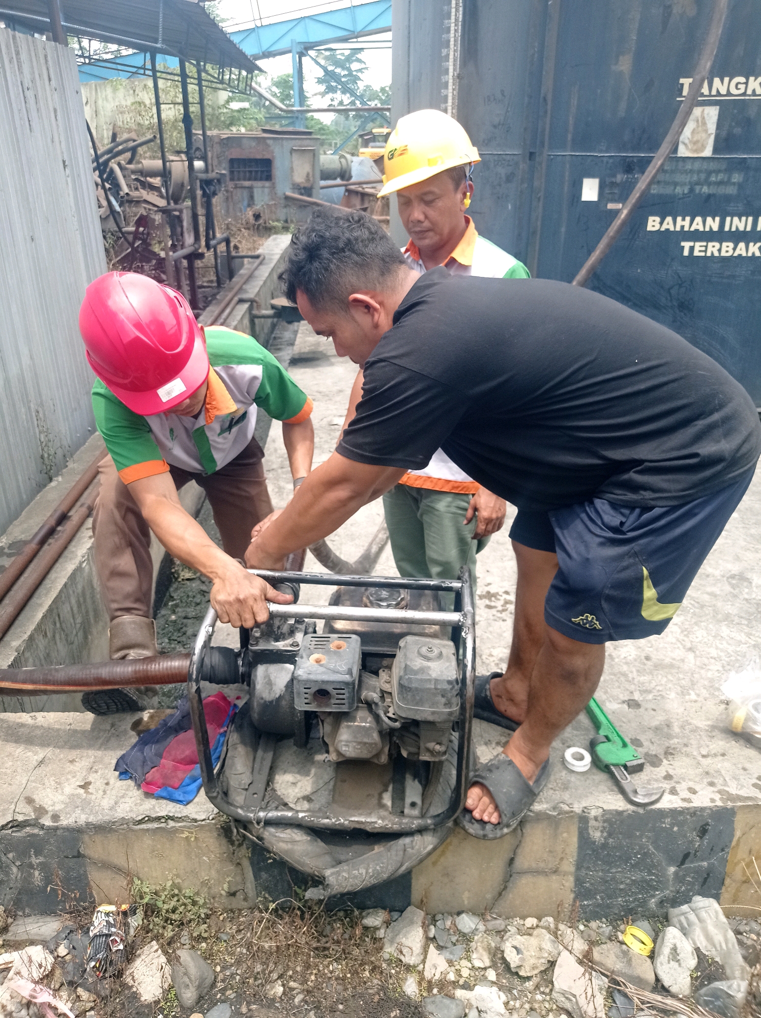 Three men are working on a portable water pump in Sumberbakti, Indonesia. One man in a red hard hat and green and orange shirt is kneeling and appears to be connecting a hose to the pump. Another man, wearing a black t-shirt and blue shorts, is also kneeling and has his hand on the pump, assisting. A third man in a yellow hard hat and green and white shirt stands behind them, observing. The scene is outdoors, with a concrete base and some debris on the ground, including rocks and vegetation. A large, dark blue tank with writing on it is visible in the background. There are also pipes and metal structures, suggesting an industrial or maintenance area. A green pipe wrench and a white sealing tape are on the ground near the pump.