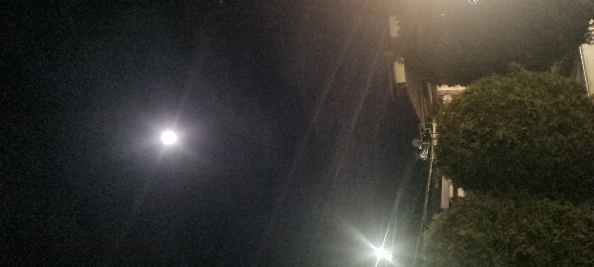 The image shows a night scene outdoors. In the dark sky is a bright, circular light source, possibly the moon. In the lower right corner are green, rounded bushes. A building with visible windows and a satellite dish are located behind the bushes. In the lower center is a bright street light. The location is likely a residential area.