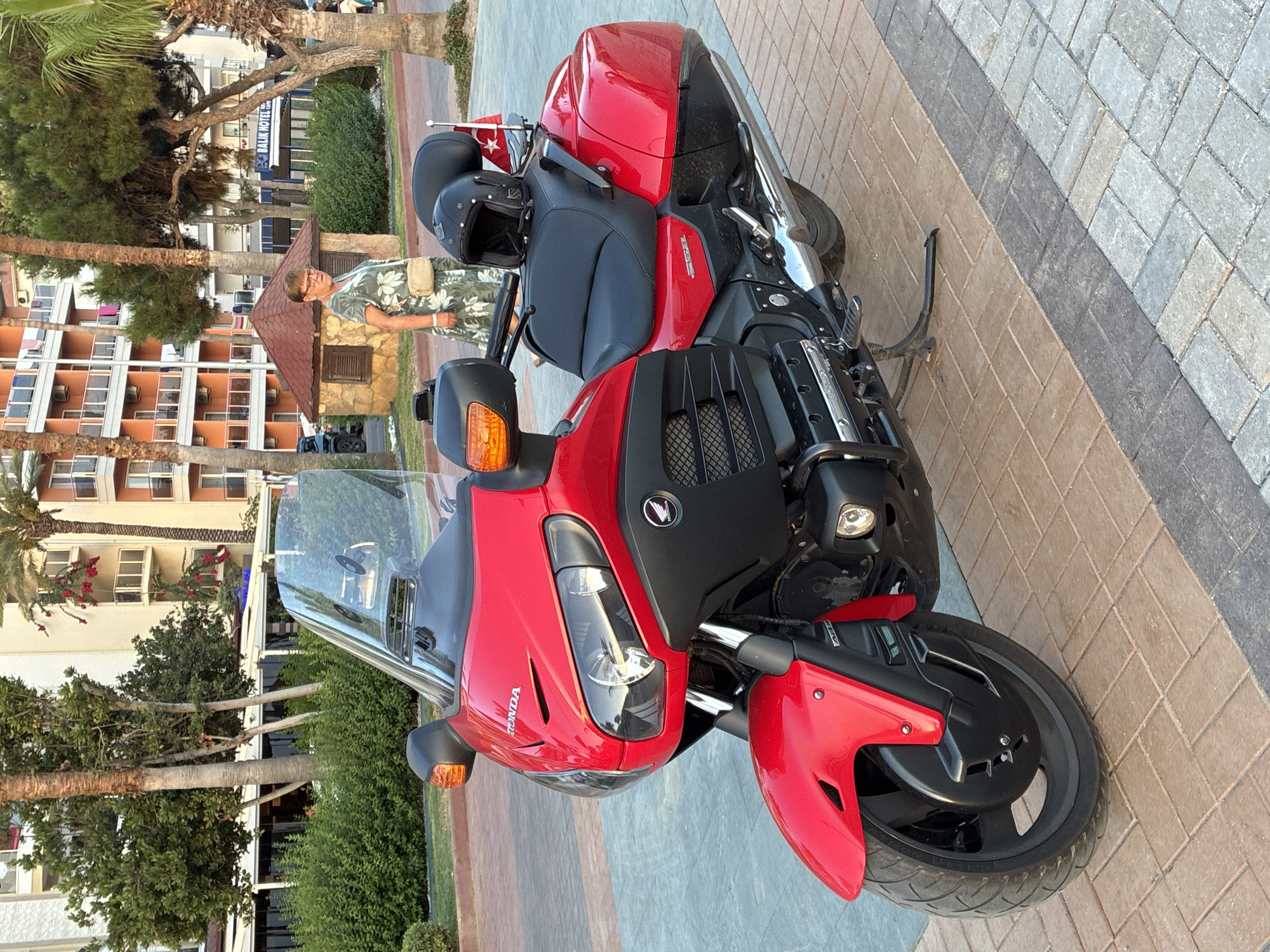 In Alanya, Turkey, a red Honda Gold Wing motorcycle is parked on a patterned stone pathway. The motorcycle is angled slightly to the right, with its front wheel closer to the viewer. A black helmet rests on the passenger seat of the motorcycle, and a small flag with a white star on a red background is attached to the rear of the bike. In the background, a woman in a floral dress walks past a small, rustic building with a red roof. Behind her are palm trees and the facade of a multi-story hotel with orange and cream-colored walls and balconies. The scene is illuminated by natural sunlight, casting subtle shadows on the ground.