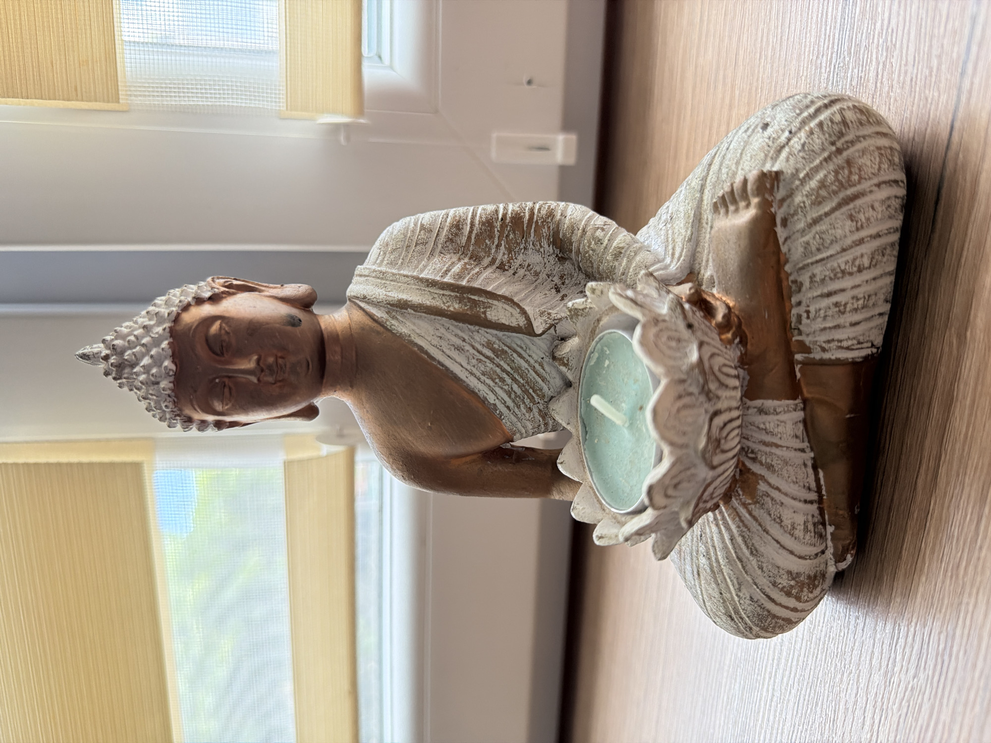 A medium shot captures a small, seated Buddha statue holding a lotus-shaped tealight holder with a pale green candle inside. The statue is made of a material that appears to be resin or wood, with a bronze-like finish on the skin and a distressed white finish on the robe and base. The Buddha is in a meditative pose, with eyes closed and hands cradling the candle holder. The background consists of a window with horizontal blinds, allowing diffused natural light to enter. The blinds are a light beige color. The statue is placed on a wooden surface, likely a table or shelf, which has a visible wood grain pattern. The overall impression is one of tranquility and peace. There is no indication of the city of Pazardzhik, Bulgaria, in the image.