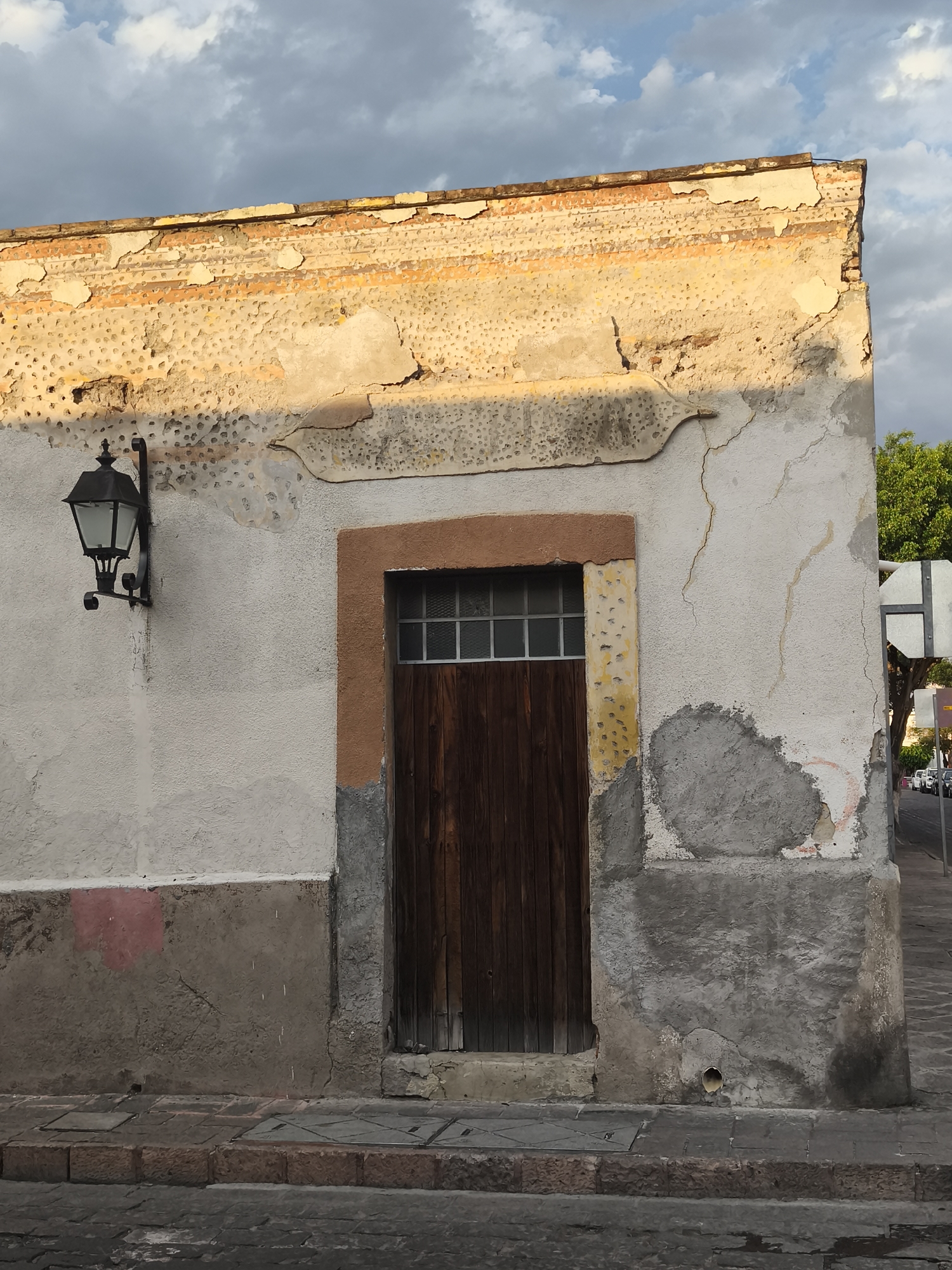 The image shows a building facade in Querétaro, Mexico. The building has a weathered appearance with peeling paint and visible damage. A dark brown wooden door is centered in the frame, with a small window above. The door is surrounded by a brown frame, with some yellow discoloration. A black metal lantern is mounted on the wall to the left of the door. The wall is off-white with a darker, grey base. The upper portion of the wall is lit by sunlight. The sky is visible in the upper right corner, with blue and white clouds. A street sign, trees, and parked cars can be seen in the background to the right of the building. The sidewalk in front of the building is made of stone.