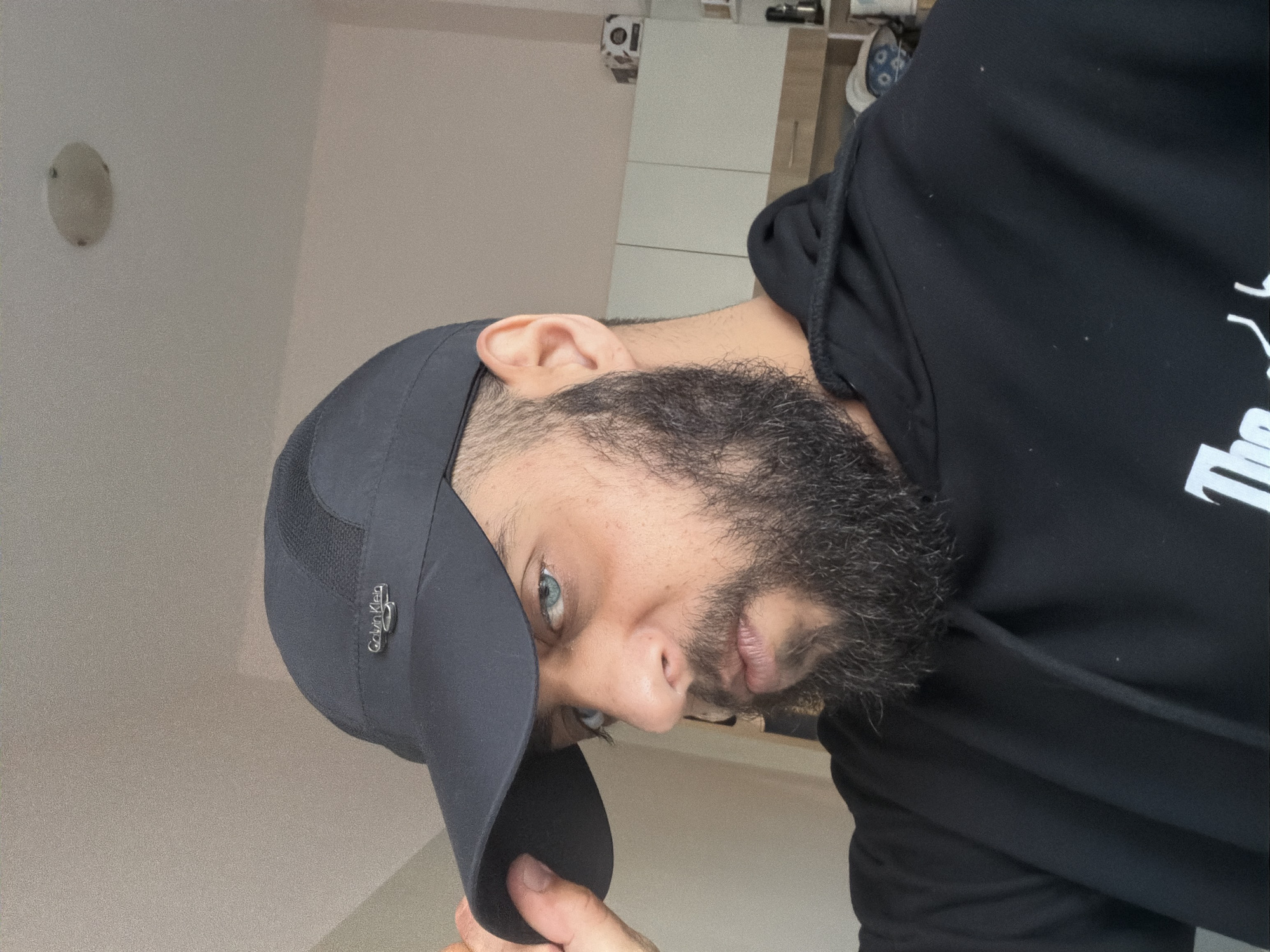 A man is wearing a black Calvin Klein baseball cap and a black hoodie. The background is a white wall and a glimpse of an interior. The location and city are unknown.