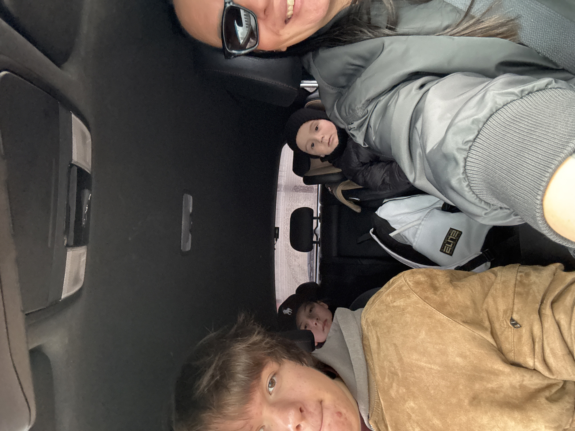 The image shows four individuals inside a vehicle. An adult woman with sunglasses is visible in the top right. A young child wearing a black beanie is positioned in a car seat. Another individual with brown hair and a tan jacket is in the foreground. A fourth individual can be seen behind him. A white "Elite" branded backpack sits behind the car seat. The interior of the vehicle appears dark, except for the light entering through the back window. The location is stated as being in the city of Chaikovsky, Russia.