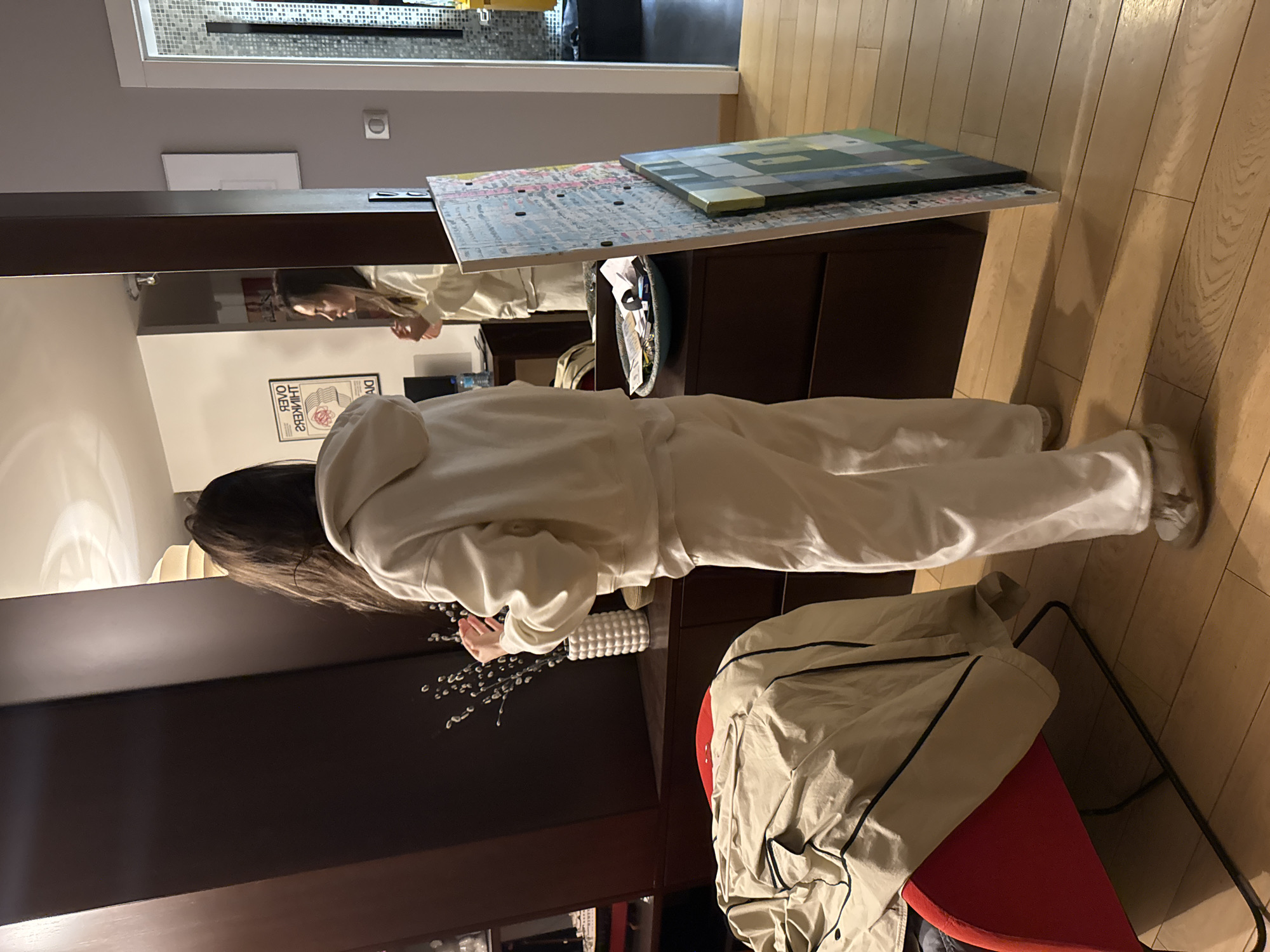 The image captures an indoor scene, likely a modern residential space in Warsaw, Poland. A person, viewed primarily from behind, stands facing a dark brown dresser or console table. They are dressed in a light-colored (possibly beige or off-white) hooded tracksuit, comprising a hoodie and matching sweatpants, paired with light-toned sneakers. Their long, dark hair cascades over their shoulders. Their hands are positioned near a white, textured vase or decorative object holding several delicate stems with small, white, fluffy catkins.
On the dresser, two large, flat items are displayed. One is a piece of abstract art or possibly a large puzzle, featuring a mosaic of geometric shapes in shades of green, blue, yellow, and gray. Adjacent to it, another flat object appears to have printed text or intricate patterns, though it is too small to decipher clearly.
A large mirror on the wall reflects parts of the room, including the person's profile as they look downwards or at the items on the dresser. In the mirror's reflection, a framed poster is visible on the wall, featuring vertically oriented, reversed text, partially decipherable as "THIKEB2" and "OAEB". A plastic water bottle can also be seen reflected on the dresser.
The room features light-colored wooden floorboards and light grey walls. In the lower right foreground, a red chair is partially visible, draped with a light beige or khaki jacket that has dark trim. The space is brightly lit, suggesting either ample natural light or strong artificial illumination. The person's posture and neutral expression suggest a calm, observant activity, possibly arranging the decorative items.