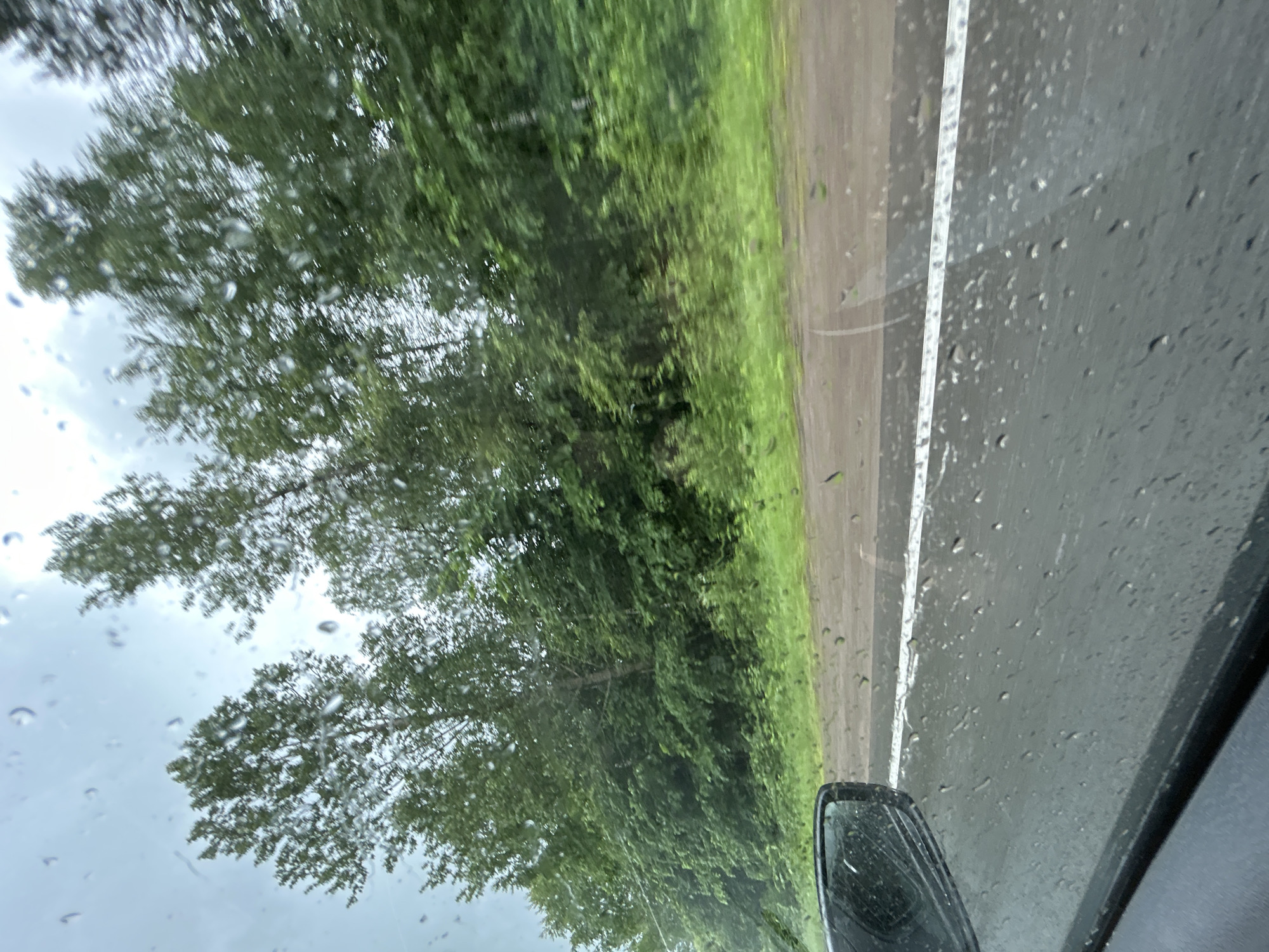 The image shows a view from inside a vehicle looking out through a rain-streaked windshield. The scene depicts a road with a white line marking the lane, bordered by lush green trees and vegetation. The sky is overcast, suggesting a rainy day. A side-view mirror of the vehicle is visible in the lower left corner of the frame. The location is identified as the city of Kulmas, Russia.