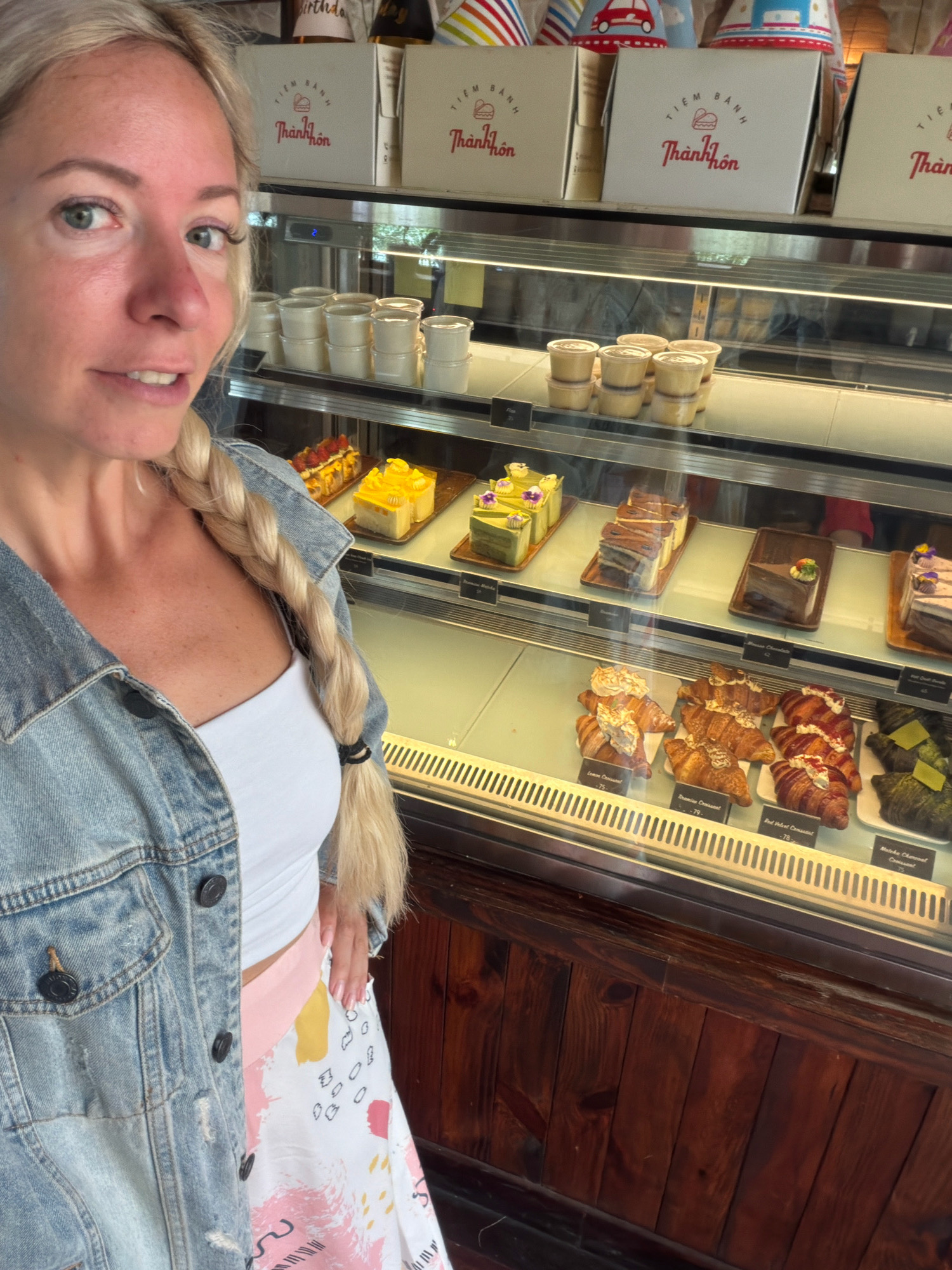 A woman with blonde hair braided down her back poses for a selfie in front of a display case filled with pastries. She is wearing a denim jacket over a white top and a skirt with a colorful abstract print. The display case features various cakes, croissants, and what appear to be small cups of dessert. In the background, boxes with Vietnamese writing are visible, along with striped party hats. The establishment appears to be a bakery or pastry shop. The signage suggests the location is a "Tiệm Bánh Thành Hôn," which translates to "Thành Hôn Bakery" in Da Lat, Vietnam.