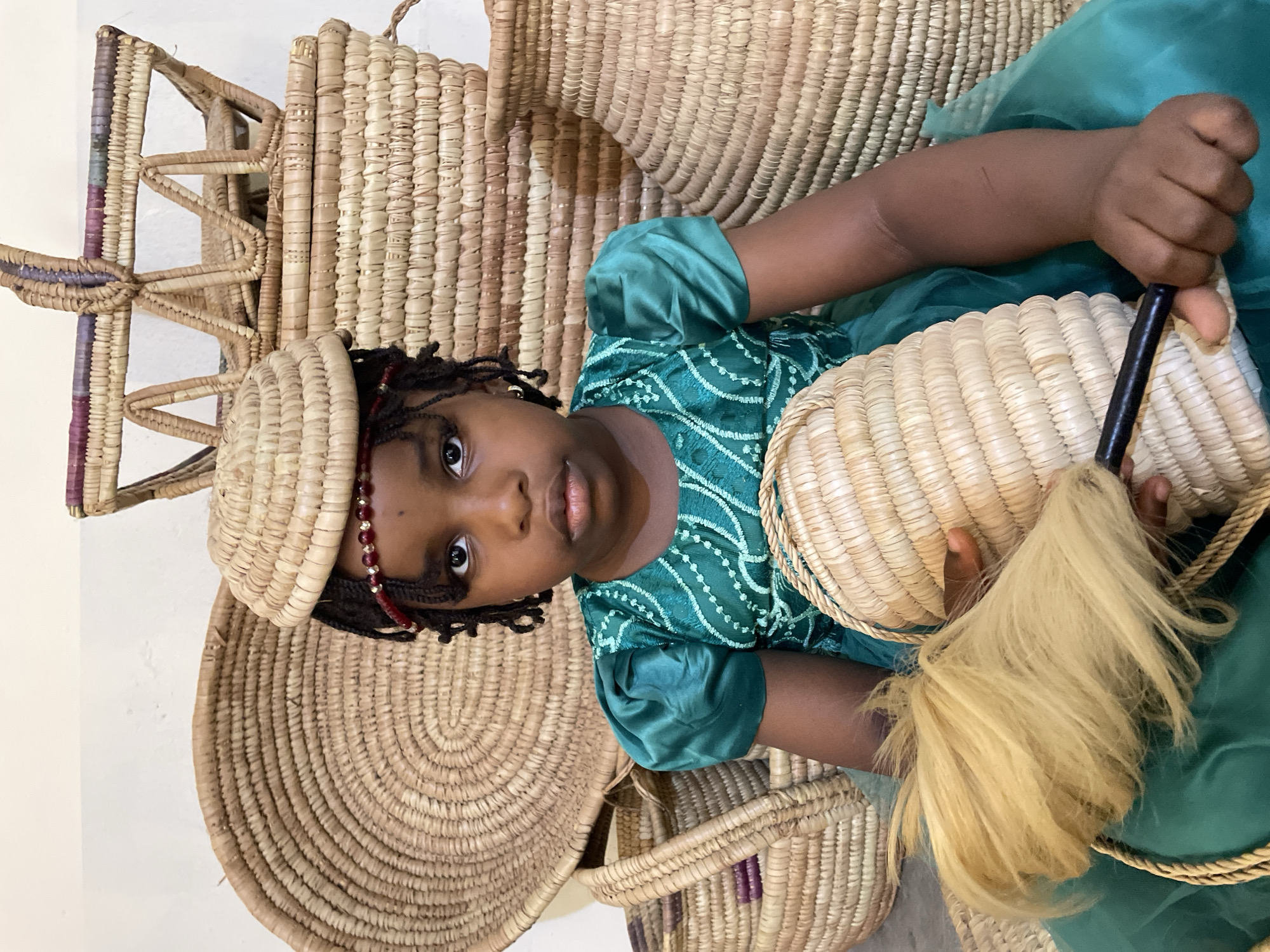 A young girl wearing a green dress and a woven hat is surrounded by various woven baskets. She is holding a basket with blonde fibers emerging from it. The location is implied to be Jos, Nigeria.