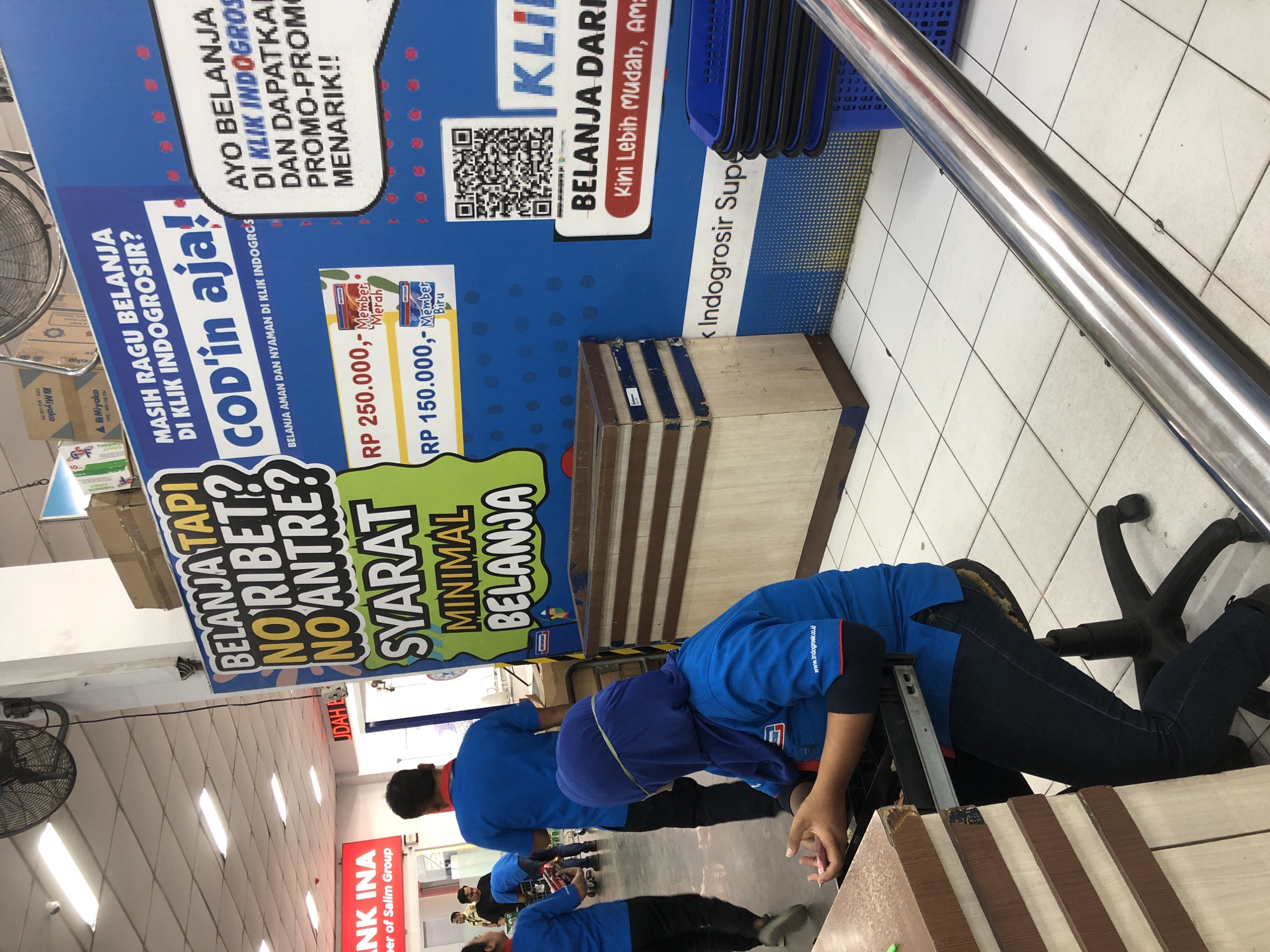 The scene is an indoor retail environment in the City of Medan, Indonesia. In the foreground, several people wearing blue shirts are positioned around what appears to be a countertop. One person is sitting on an office chair, and another person is bending over, possibly working at the counter. In the background, an advertisement display with text written in Indonesian is visible. The display includes prices for memberships and promotes online shopping via Klik Indogrosir. The floor is tiled, and there are some storage boxes and shopping baskets visible. In the distance, there is an ATM visible from "BANK INA." A ceiling fan is located on the left.