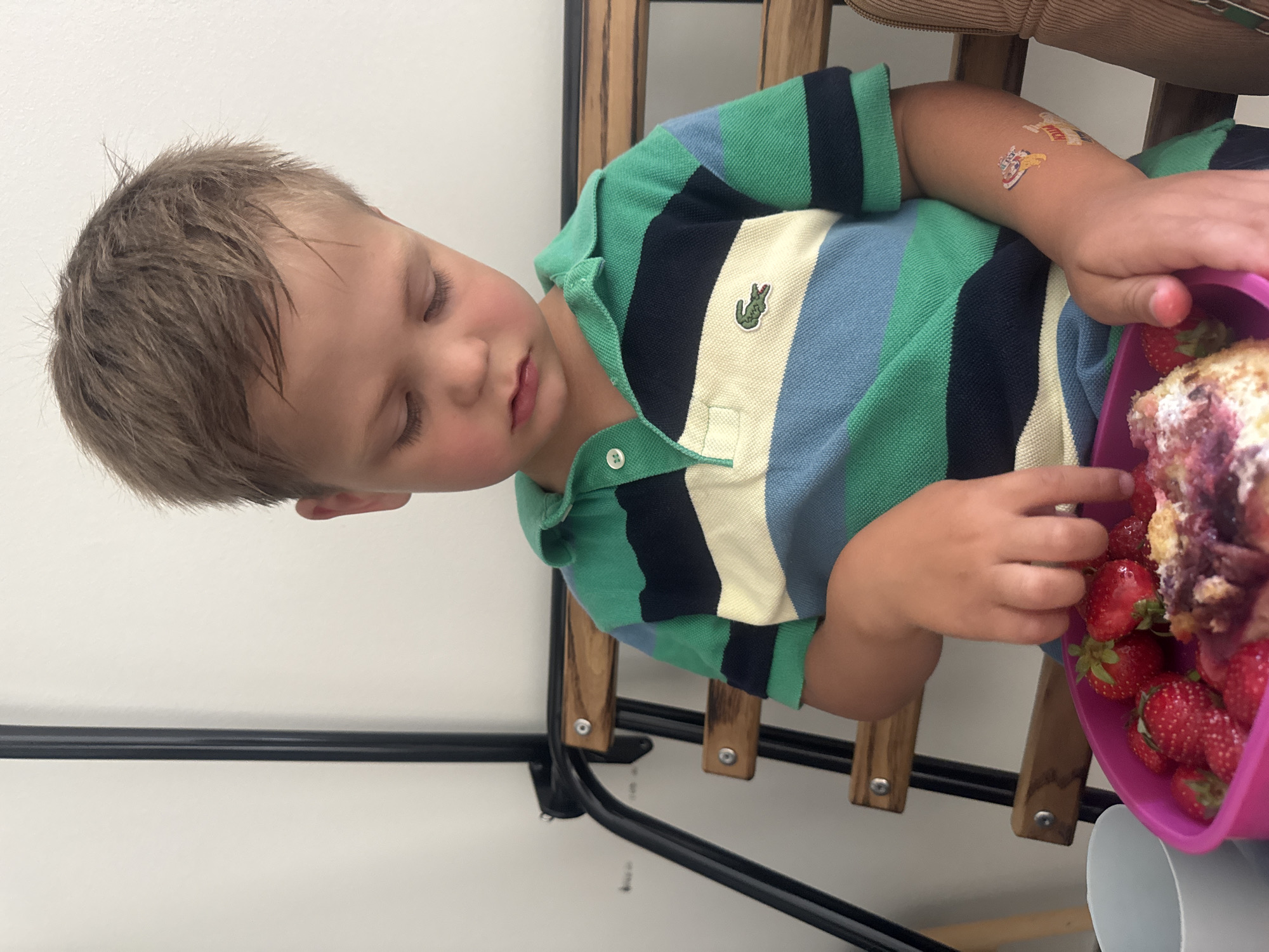 The image shows a young boy sitting on a wooden bench, looking down at a pink bowl of strawberries and dessert. The boy is wearing a striped polo shirt with green, blue, and white stripes. There are two temporary tattoos on his right arm, one appearing to be a cartoon character and the other with text. A white cup is partially visible to the left of the bowl. The background consists of a white wall and a dark metal structure. This scene does not provide any visual cues to suggest a specific location within the city of Les Rousses, France.