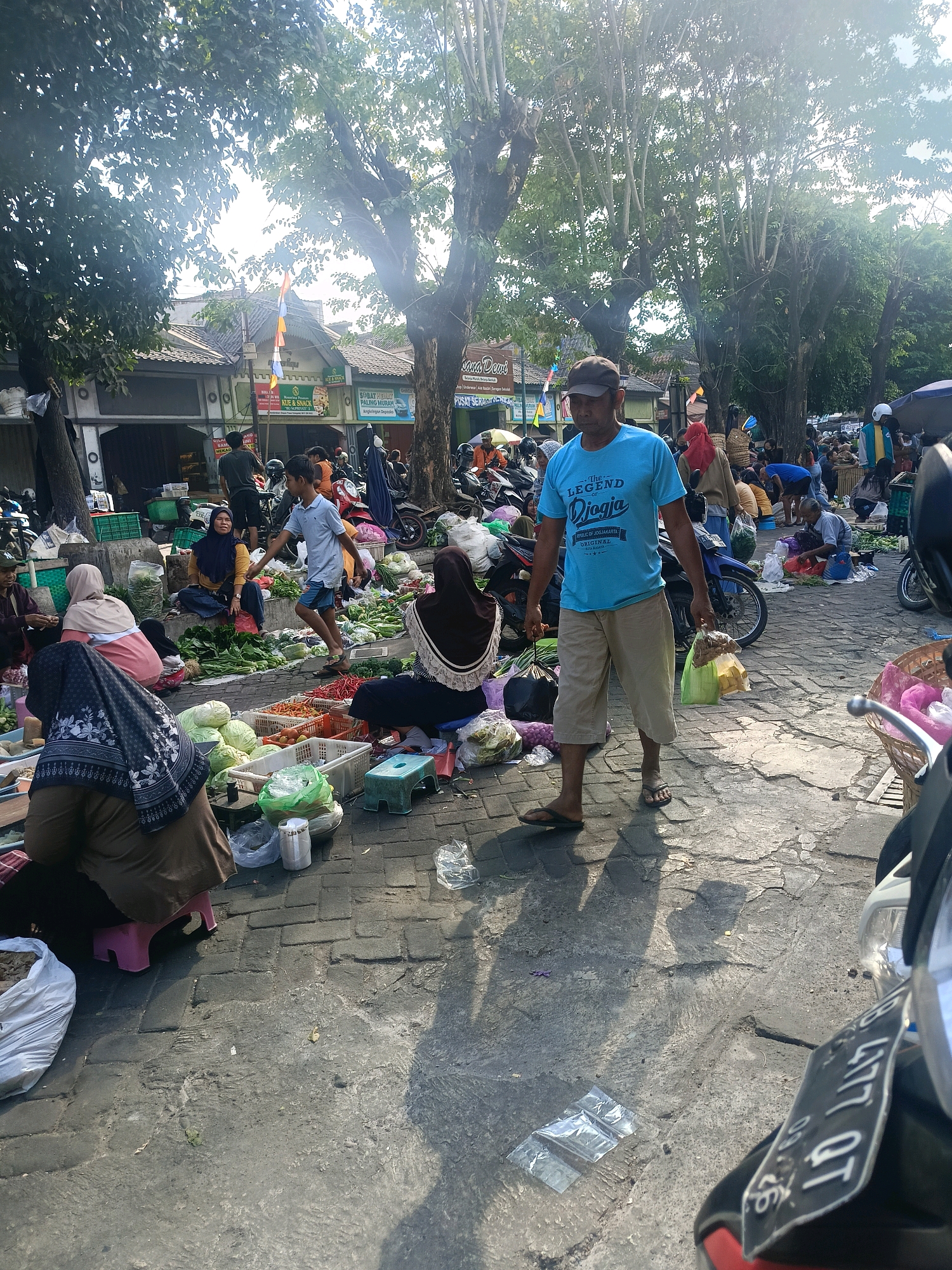 A crowded outdoor market is depicted on a cobblestone surface under a canopy of trees, suggesting a location in the city of Yogyakarta, Indonesia, likely near a local community or roadside stall. The scene is illuminated by bright sunlight, casting long shadows from the trees and people.
Several individuals are present, engaged in various activities. In the foreground, a man wearing a blue t-shirt with "The Legend of Jogja" printed on it, and tan shorts, walks with two plastic bags of goods. To his left, a woman in a dark hijab and patterned scarf is seated, partially obscured, attending to something on the ground.
Further into the market, a young boy in a light blue shirt and shorts is walking past a woman seated on the ground wearing a yellow headscarf and dark clothing. She is surrounded by piles of fresh produce, including leafy green vegetables and red chilies. Other people are visible throughout the market, some sitting and selling goods, others walking and browsing.
The ground is littered with produce, plastic bags, baskets, and some small stools. Various types of vegetables, such as cabbage and chilies, are displayed in crates and directly on the ground. In the background, there are some buildings with shopfronts, possibly stores or more permanent market stalls, and parked motorcycles. The overall impression is of a bustling, active marketplace during daylight hours.