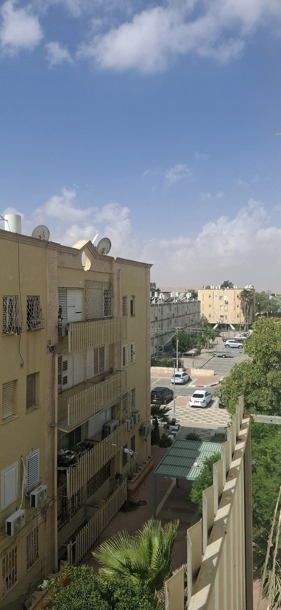 This is a vertical, eye-level, daylight photograph taken in Yerocham, Israel, showing an urban residential scene with a bright blue sky dotted with white clouds.
In the foreground, on the left, is the side of a light yellow, multi-story apartment building. This building has multiple balconies with light-colored wooden railings, some of which contain air conditioning units and satellite dishes. Several windows are visible, some with white shutters and decorative metal grates. On the ground floor, there is visible clutter on one of the balconies.
To the right of the yellow building, a paved area with parking spaces is visible. Several cars are parked, including a dark SUV and a white sedan. A small, green-roofed shelter with metal supports is also present in this area. Beyond the parking area, more apartment buildings with light-colored facades and many windows are lined up. Further in the background, a palm tree and other greenery are visible, as well as low-rise buildings and arid terrain under a hazy sky. A white trailer is parked in one of the parking spots.
A fence or railing made of light-colored vertical slats runs along the right edge of the frame, obscuring a portion of the view of the buildings and greenery to the right. The overall impression is of a sunny day in a residential neighborhood.