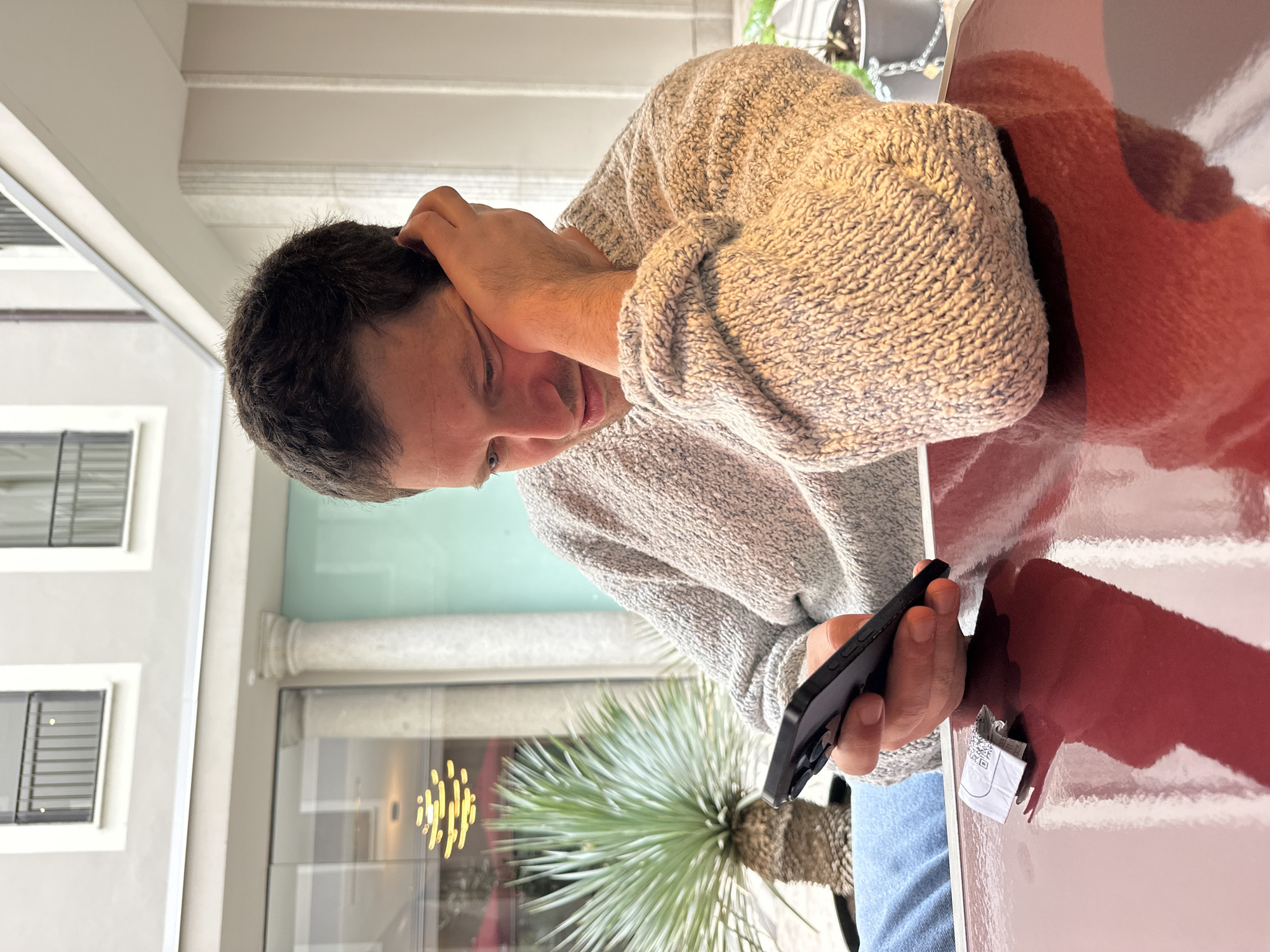 A man is sitting at a table, looking at his smartphone. He is wearing a light-colored sweater and blue jeans. His right hand is holding his head, and his left hand is holding the phone. There is a small, crumpled piece of paper next to the phone on the table. The table is a glossy reddish-brown color. Through a window, a view is seen of exterior architecture, including columns and windows, along with a green plant. The location is likely Milan, Italy.