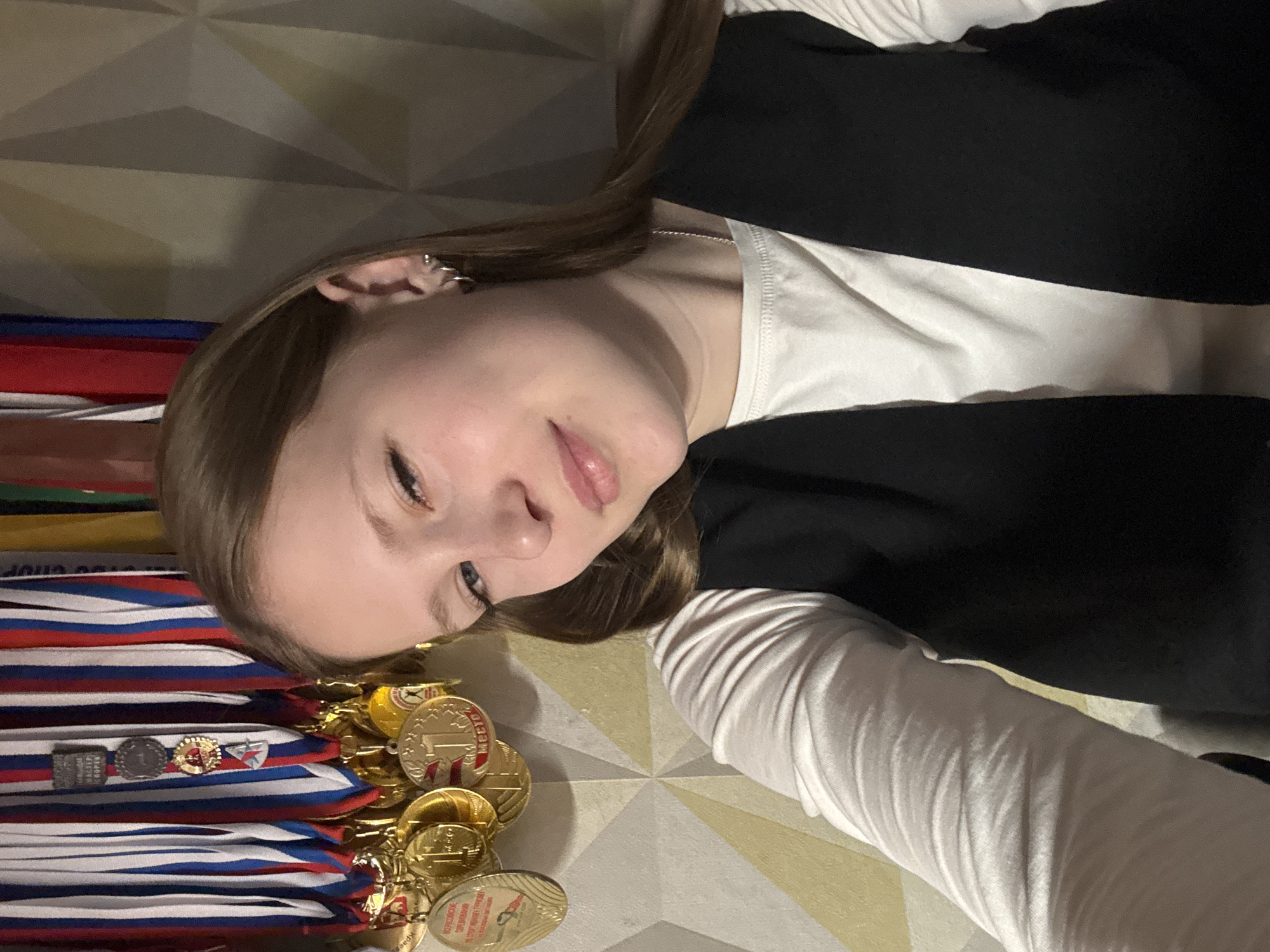 A young woman is depicted indoors, casually posing amidst a collection of numerous gold medals. She has long, light brown hair, which is parted to one side and falls over her shoulders. She wears a white long-sleeved top beneath a dark, possibly black, sleeveless garment or vest. Small hoop earrings are visible on her left ear. Her expression is relaxed, with a slight, gentle smile.
A significant number of gold medals are piled and spread around her, particularly to her left and below her. Many of these medals feature ribbons in red, white, and blue, colors that evoke the Russian national flag. The medals appear to be from various competitions, signifying athletic achievements.
Visible text on the medals includes "1 МЕСТО" (1st Place), clearly indicating top honors. One medal prominently displays "МАСТЕР СПОРТА" (Master of Sport), a high athletic title in Russia. The word "ЧАЙКОВСКИЙ" (Chaikovsky) is also discernible on a medal, aligning with the provided location context. The background features a light-colored surface with a repeating geometric, faceted pattern, suggesting a modern wall or floor design. The lighting appears to be artificial, casting some shadows and giving the scene an indoor, possibly evening, ambiance. The image captures the woman displaying her impressive sporting accomplishments.