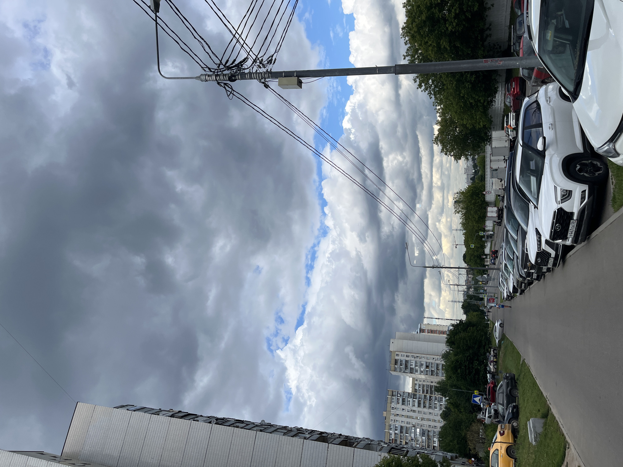 This is a vertical, eye-level shot of a street scene in Moscow, Russia, under a dramatic, cloudy sky. The foreground is dominated by the roofline of a modern, white-tiled building on the left, and a row of parked cars on the right.
The cars are mostly white SUVs and sedans, lined up along a paved road. A yellow taxi is visible further down the street, parked on the grass. Beyond the cars, a sidewalk runs alongside a grassy area with trees. In the distance, several multi-story apartment buildings are visible, characteristic of urban Russian architecture. The sky is filled with large, dark, and light gray storm clouds, with patches of blue sky peeking through. The overall mood is somewhat foreboding due to the weather.
A single person, appearing as a small figure in the distance, is walking on the sidewalk. Overhead, a complex network of electrical wires and a utility pole are prominent. There are no overtly identifiable landmarks, but the architecture and the presence of many cars suggest a residential or commercial urban area. The time of day appears to be daytime, but the heavy cloud cover makes it difficult to be precise. Some license plates are visible on the cars, with Russian characters. One visible license plate reads "Y447OA". The overall scene is of an ordinary urban day, with the weather being the most striking element.