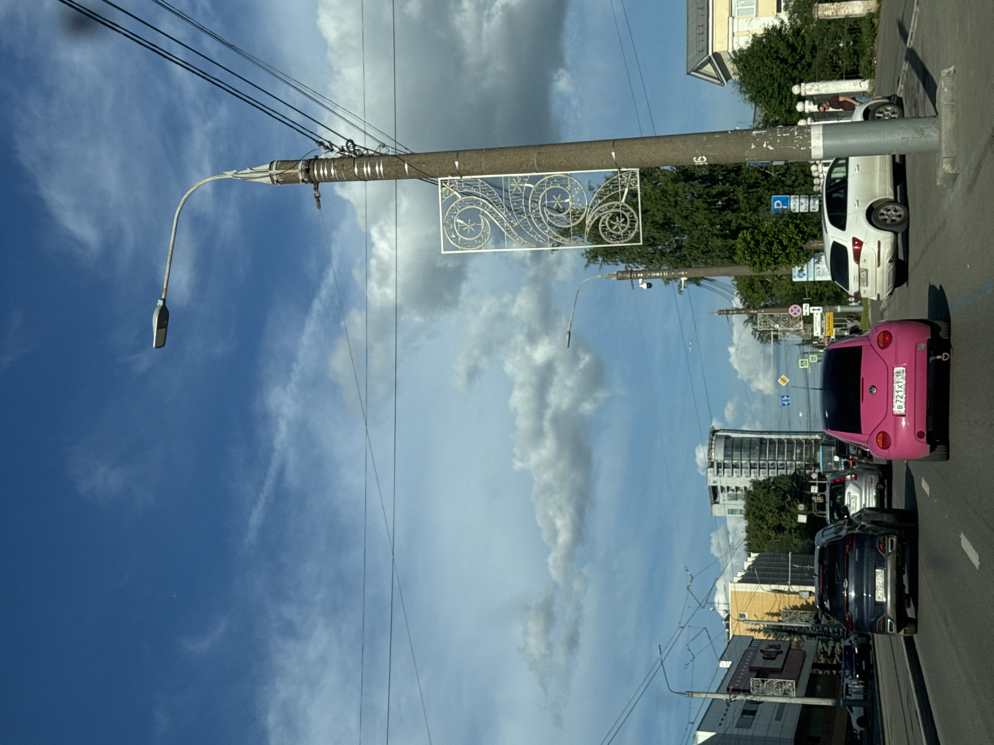 This image captures an urban street scene in Izhevsk, Russia, during what appears to be a bright day with a partly cloudy sky. The perspective is slightly elevated, looking down a road with parked and moving vehicles.
In the foreground and midground, a large concrete utility pole stands prominently on the left side of the road. It supports a street lamp that curves downwards, overhead power lines, and a decorative white metal panel featuring intricate, swirling patterns.
On the right side of the road, several cars are visible. A pink, compact car, possibly a Volkswagen Beetle, is closest to the viewer, facing away. Its Russian license plate, "B 721 XT 18," is clearly readable. Behind it, a dark blue SUV with a partially visible license plate "P327MY 177" is in traffic. Further down the street, a white SUV is parked next to the utility pole, with a person partially visible near its rear wheel. The white SUV also has a partially visible license plate, "P224OY 72". A blue parking sign with a white 'P' is located near the white SUV.
The street is lined with various buildings, including a multi-story structure with yellow and white facades, and taller, more modern buildings in the distance. Lush green trees and bushes are scattered along the roadside, separating the pavement from some of the buildings. The sky is a mix of clear blue and white cumulus clouds. No specific event is taking place; the scene depicts everyday city life and traffic.