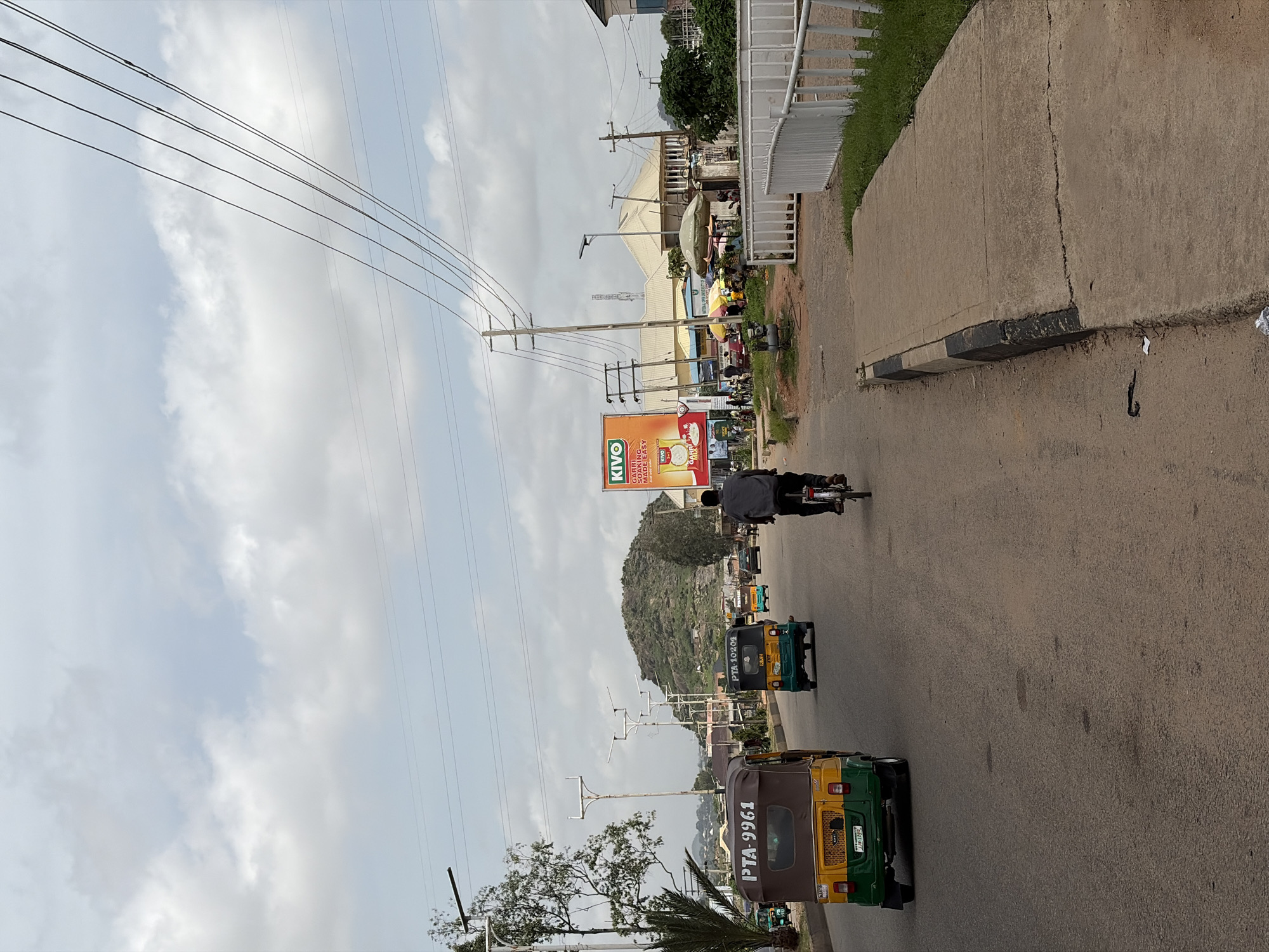 This is a photograph of a street scene in Gura Top, Nigeria. The image is oriented vertically. The foreground shows a paved road with several auto-rickshaws (also known as Keke Napep or Marwa) driving in the same direction. The auto-rickshaws are colorful, with two of them clearly displaying license plates: one reads "PTA-10209" and the other "PTA-9961". A person is riding a bicycle in the background.
The middle ground features buildings, some with awnings, and a large billboard for "KIVO" soap. Power lines stretch across the scene, supported by utility poles. In the background, a lush, green hillside rises under a cloudy sky.
The setting appears to be a busy street, likely during daylight hours given the illumination. The weather seems to be overcast or partly cloudy. The overall impression is one of a typical urban or semi-urban environment in Nigeria.