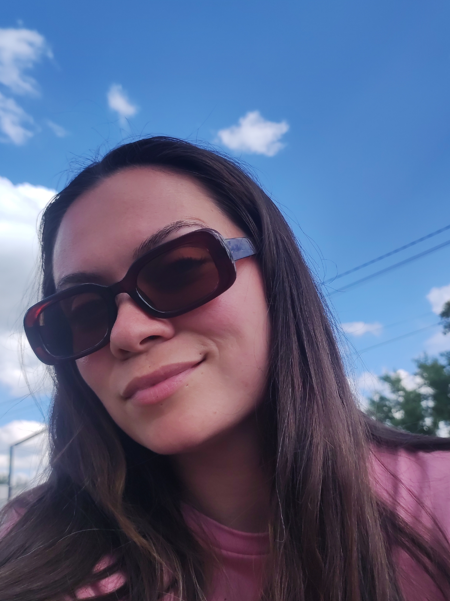 A young woman with long, dark brown hair is looking up at the camera, her face partially obscured by large, dark sunglasses with a tortoiseshell pattern. She is wearing a bright pink shirt. Her skin has a warm, rosy glow, suggesting a sunny day. The background is a bright blue sky with scattered white, fluffy clouds. The angle of the photo, with the sky prominently featured, suggests it was taken outdoors during the daytime. The overall impression is casual and relaxed, perhaps a selfie taken on a pleasant day. There are no visible text elements in the image.