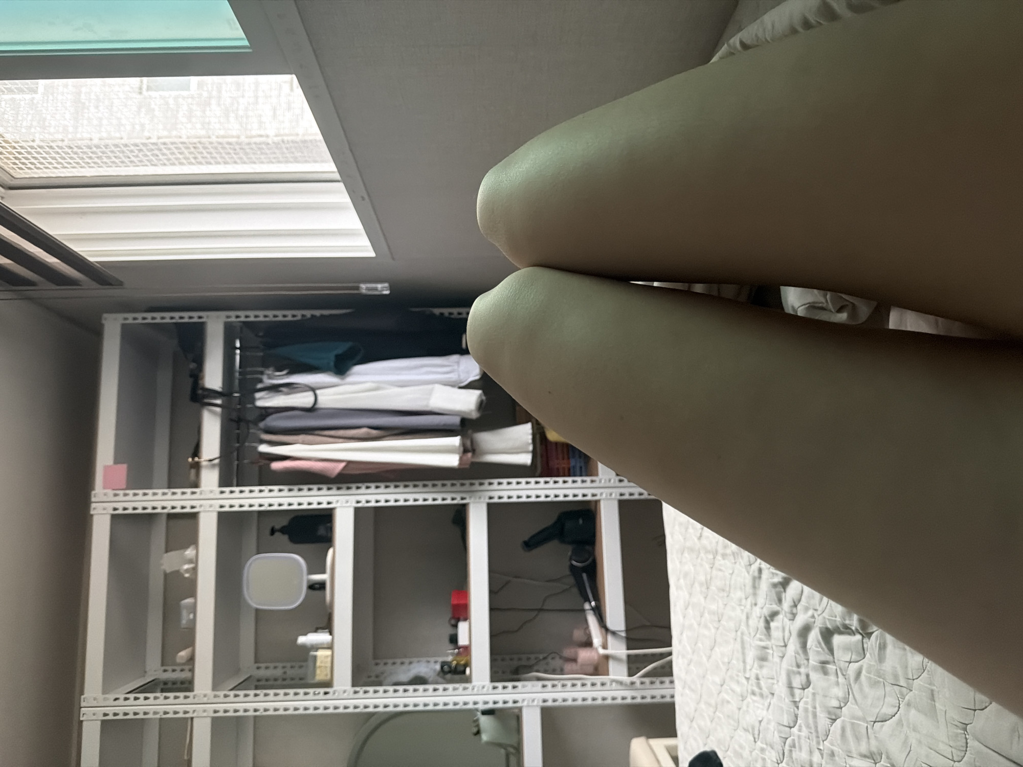 The image depicts a close-up view of a person's thighs and knees, resting on a quilted bed. In the background, a white metal shelving unit is visible, displaying various items including clothes on hangers, a mirror, bottles, and what appears to be a hairdryer. Adjacent to the shelving unit is a window with blinds. The overall setting appears to be a bedroom or living space within a residential building in Dongducheon-si, South Korea. The lighting suggests it is daytime.