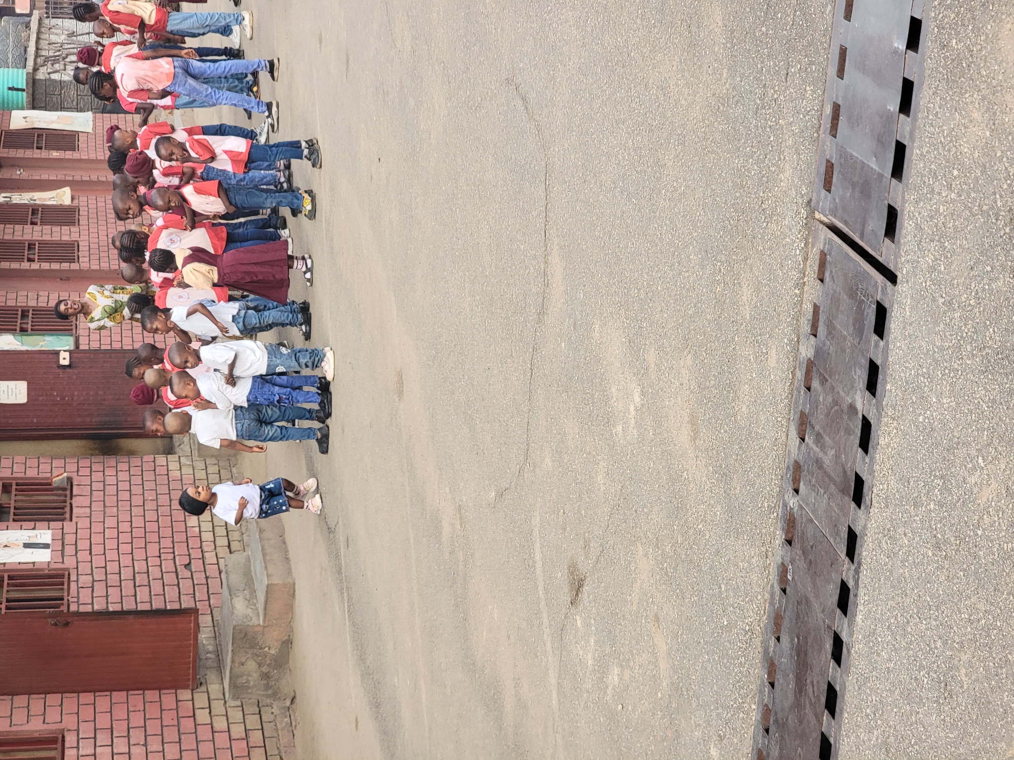 In Maiburiji, Nigeria, a group of children in uniforms stand in a line along a paved street next to a brick building. The children are wearing varying shades of white shirts with blue denim jeans or skirts. Some have red or pink vests. A woman in a colorful patterned top stands near the middle of the line. A drainage ditch with a metal grate runs along the side of the road.