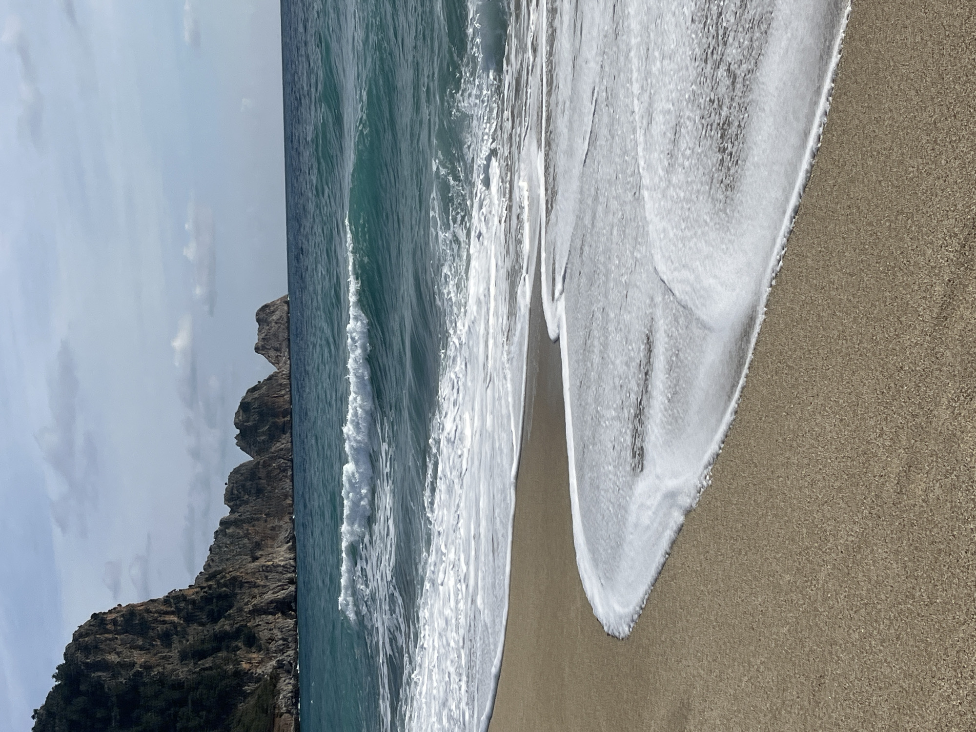 This image displays a coastal scene near Alanya, Turkey. The foreground consists of light tan-colored sand, partially wet, with white foam from receding waves forming distinct, curvilinear patterns. The midground is dominated by the turquoise-blue Mediterranean Sea, featuring multiple breaking waves with white crests and foam. In the background, to the left, a substantial, dark, rugged cliff face rises directly from the sea, exhibiting sparse green vegetation near its summit. Several distinct, smaller rock formations or islets are visible further offshore in the center-right background. The sky above is partly cloudy with light grey formations. No human figures, animals, or man-made structures are present within the frame.