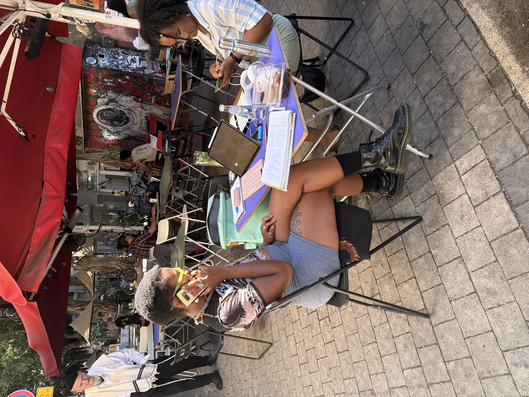 The image captures a scene in Tel-Aviv, Israel, featuring two individuals seated at a makeshift workspace along a brick-paved pedestrian area. One person, clad in a striped shirt, is focused on their phone, while the other, sporting shorts and a patterned top, is engaged in a phone call, holding a yellow-cased phone to their ear. A table between them is laden with a laptop, notebook, food containers, water bottle, and various other personal items. The area behind them is shaded by a red awning and appears to be part of an outdoor market or craft fair, with vendors and passersby visible in the background.