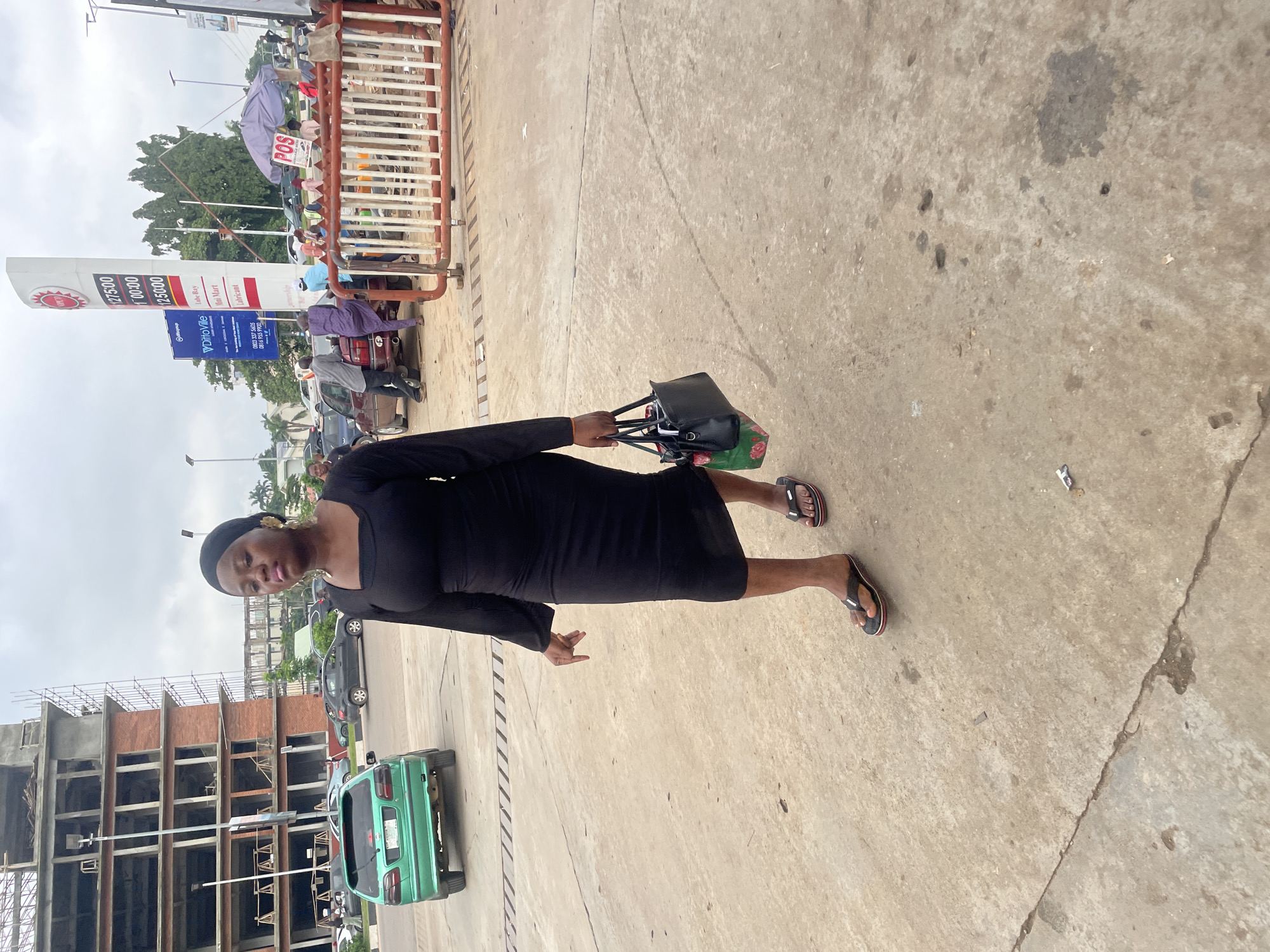 A woman in a black dress and headscarf walks on a concrete surface in Abuja, Nigeria. She carries a black handbag with a floral bag underneath. In the background, a multi-story building under construction and a gas station with a price board are visible. Several cars are parked or driving on the road. Other people are also present, some near the gas station and others behind a red barrier. The sky is overcast.