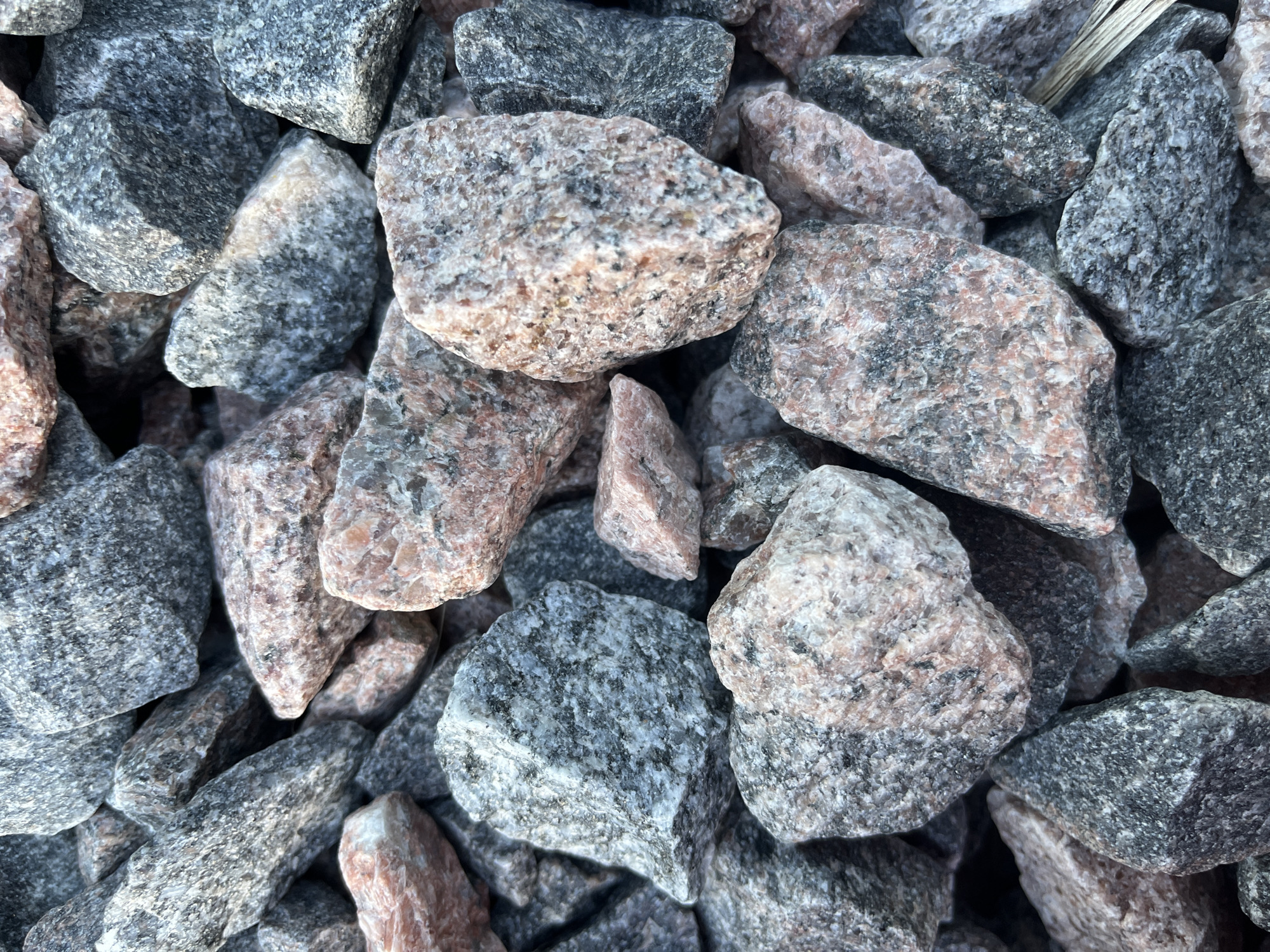A close-up, overhead shot shows a pile of granite gravel. The stones are angular and vary in size, with most measuring between 1 to 3 inches. The gravel is a mix of gray, pink, and black speckled pieces. The lighting suggests it is daytime. There are no people or animals visible in the image. The environment is a generic outdoor setting, possibly a construction site or landscaping project. The specific location of Il'ino, Russia, cannot be determined from the visual information provided in the image.