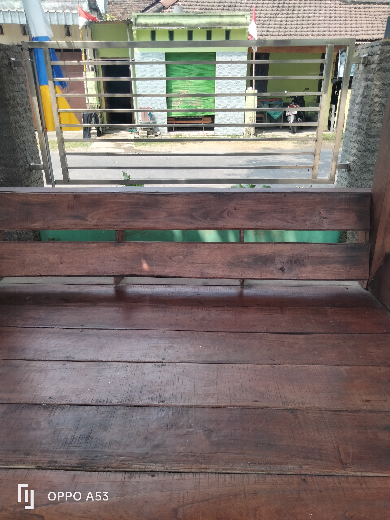 The image shows a close-up view of a wooden bench, with a metal fence and buildings in the background. The bench is made of dark-stained wood planks. Through the fence, there are several buildings with tiled roofs. One building has green rolling shutters and another has a red and white flag displayed. A motorcycle is visible parked in front of one of the buildings. The scene appears to be outdoors, possibly in a residential area. The "OPPO A53" watermark is present in the bottom right corner of the image, indicating the phone model used to capture the photograph. The image was taken in Jati, Indonesia.