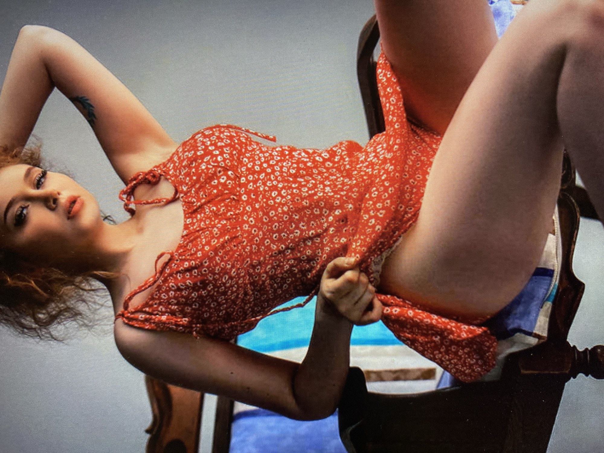 A young woman with auburn curly hair is seated in a wooden chair. She is wearing a red floral dress with thin straps. Her right arm is raised behind her head, and her left hand is pulling up the hem of her dress. The chair has a blue and white striped cushion. The background is a plain gray wall. The image is taken from a medium-close-up angle, focusing on the woman's upper body and legs.