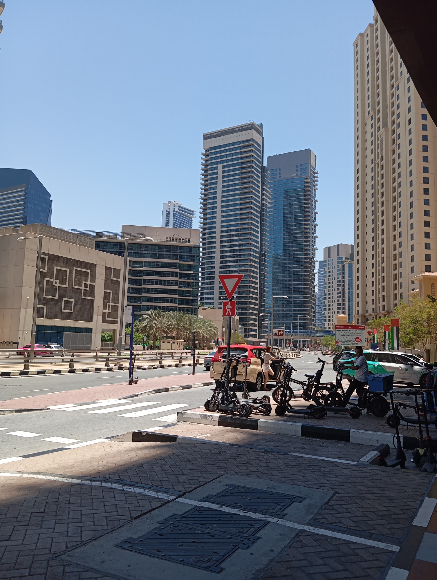 The image captures a street scene in Dubai, United Arab Emirates, likely in a modern urban district. Tall skyscrapers with glass and concrete facades dominate the background under a clear blue sky. In the foreground, a street with clear lane markings and a pedestrian crossing is visible. Several electric scooters are parked along the sidewalk, some of which are being used or attended to by people. A few vehicles, including a red car and a white SUV, are present on the road. The architecture suggests a contemporary city environment with a focus on urban development and transportation.