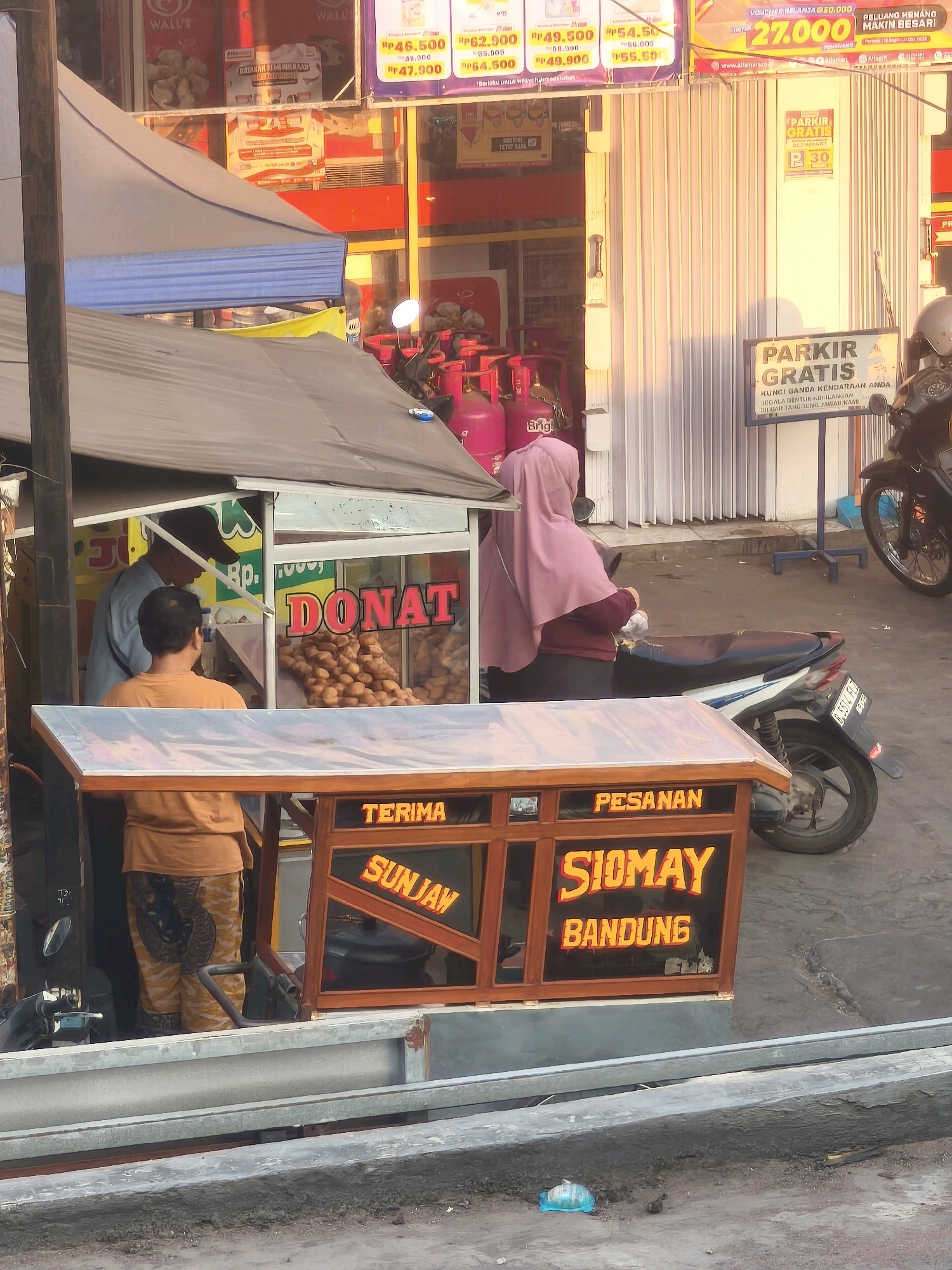 In an outdoor urban environment, a food cart vendor is serving customers. The cart, constructed of wood and glass, displays "DONAT" (donuts) and "SIOMAY BANDUNG" (a type of Indonesian dumpling). A vendor, wearing a light blue shirt and a dark cap, stands behind the counter. A customer, dressed in an orange t-shirt and patterned pants, faces the cart. Adjacent to the food cart, a motorcycle is parked. In the background, a store with red and yellow signage is visible, featuring promotions such as "PARKIR GRATIS" (free parking). The scene is illuminated by the warm light of what appears to be late afternoon sun.