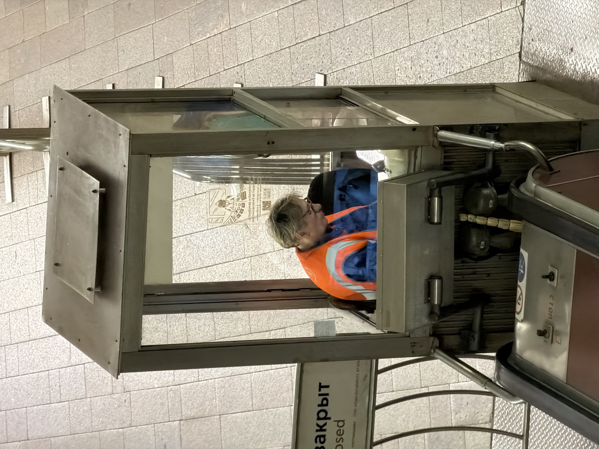 The image shows a woman inside a closed escalator operator booth. The booth is made of metal and glass, with a sign in Russian that translates to "closed". The woman is wearing a blue jacket and an orange safety vest. The escalator is also metal and red. The image was likely taken in a metro station in Moscow, Russia, based on the Russian writing on the sign and the style of the station.