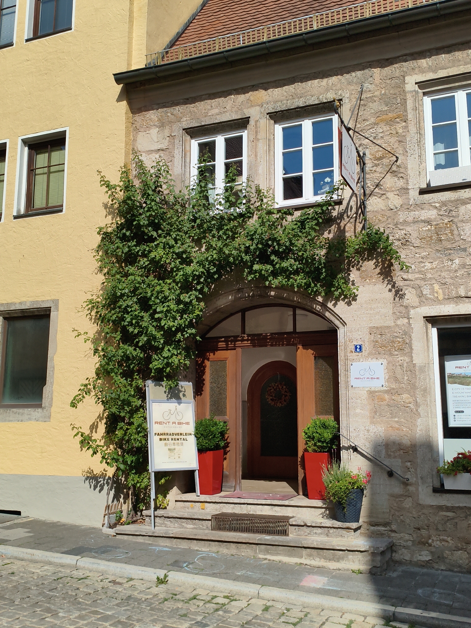 The image depicts the entrance to a bike rental shop in Rothenburg ob der Tauber, Germany. The shop is located in a building with a yellow facade on the left and a stone facade on the right, characteristic of the historic architecture of the town. A vibrant green vine climbs up the stone wall, framing the arched doorway. The doorway itself is made of wood and leads into a dimly lit interior where a wreath hangs.
Outside the entrance, a sign displays "RENT A BIKE ROTHENBURG" with a bicycle graphic and information in German and Japanese. Next to the sign, a bright red planter holds a small green bush. To the right of the entrance, a set of stone steps leads up to the door. A dark gray planter with a flowering plant sits on the steps. Further to the right, a white planter with red flowers is visible near a window. The street in front of the shop is paved with cobblestones. The scene is bathed in sunlight, casting shadows on the building and the street.