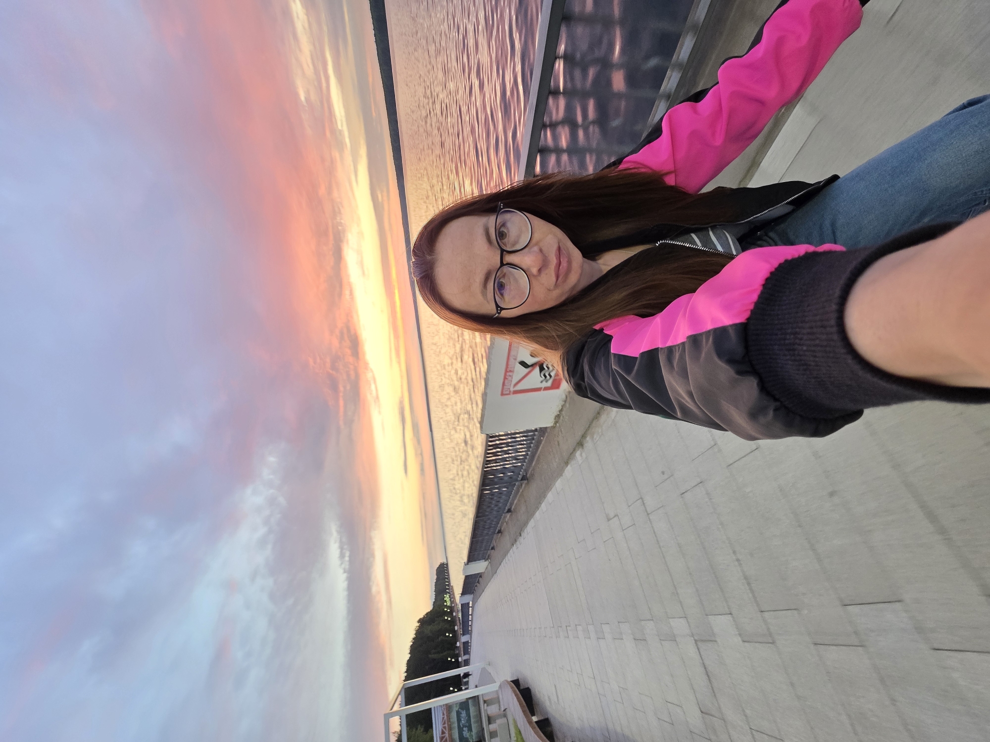 A woman with long brown hair and glasses takes a selfie on a paved promenade overlooking a wide body of water at sunset. The sky is filled with vibrant colors of pink, orange, and purple as clouds drift by. The promenade is lined with a metal fence and benches. Trees and buildings are visible in the distance along the shore. The woman is wearing a black jacket with bright pink sleeves and blue jeans. The scene appears to be along the Kama River embankment in Chaikovsky, Russia.