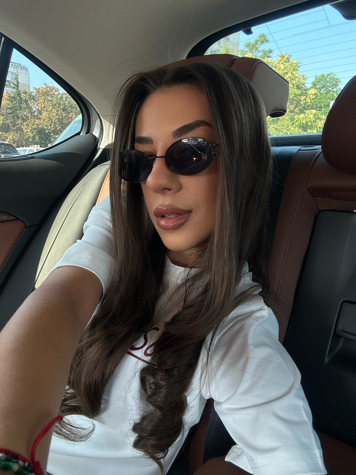 In the city of Sofia, Bulgaria, a young woman with long, dark brown hair and dark-rimmed sunglasses is sitting in the back seat of a car. She is wearing a white t-shirt. Her left arm is extended towards the camera, and she is wearing a red bracelet on her wrist. The interior of the car has dark brown leather seats and a grey headliner. Through the car windows, trees and buildings are visible, indicating an urban or suburban environment. The lighting suggests it is daytime.