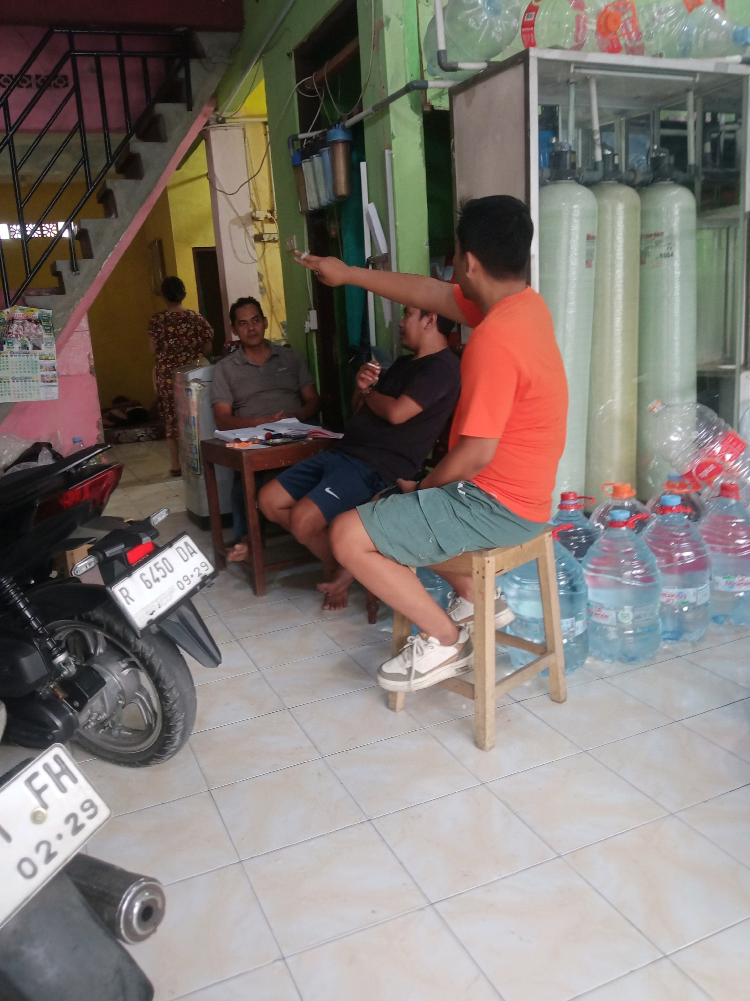 The image depicts a scene in Grendeng, Indonesia, likely a shop or a home with a commercial operation. In the foreground, a motorcycle is parked, with its license plate clearly visible, reading "R 6450 DA 09-29". To the right, three men are present. One man, wearing an orange t-shirt and green shorts, is seated on a wooden stool and appears to be gesturing. Another man, in a grey polo shirt, sits at a wooden table with papers. A third man, wearing a black t-shirt, is also seated. Behind them, a water filtration system is visible, with several large water bottles stacked around it. The environment suggests a utilitarian space, possibly a water refilling station, with elements of a residential setting such as a staircase in the background.