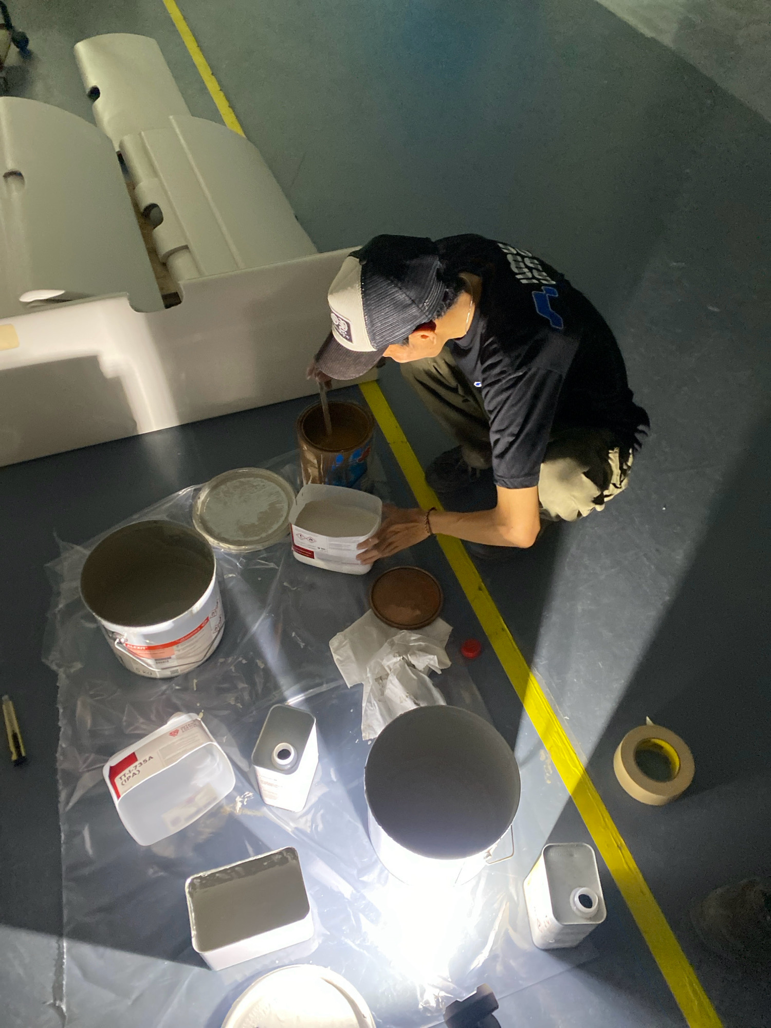 In Batam, Indonesia, a person wearing a black shirt, a baseball cap, and olive-colored pants kneels on a gray floor. The person is in the process of mixing a light brown substance with a long tool in a dark brown, open can. The surrounding area is covered with a clear plastic sheet, on which are various containers of materials. These include a large white bucket with a red and white label, a flat gray lid, a white plastic container with a red and white label, a small white container, and a rectangular white container with gray contents. There is also a round, shallow brown dish, a roll of masking tape, and a yellow line marking on the floor. In the background, a large, white, curved object, possibly part of a structure or vehicle, is visible. The scene appears to be an indoor workspace.
