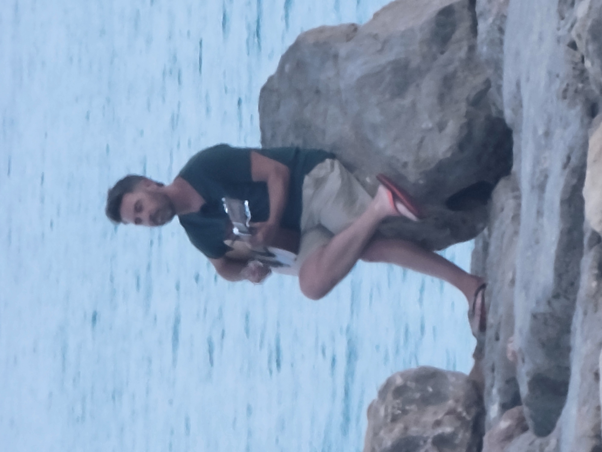 A man is sitting on a large rock by the sea, playing a guitar. He is wearing a dark green t-shirt and khaki shorts. The sea is calm and a light blue. The man has short dark hair and a beard. His right leg is crossed over his left, and he is wearing red flip-flops on his feet. The rock he is sitting on is large and grey, with a rough texture. The foreground shows more rocks, suggesting a coastal path or area. The image appears to be taken in Quarteira, Portugal, given the coastal setting.