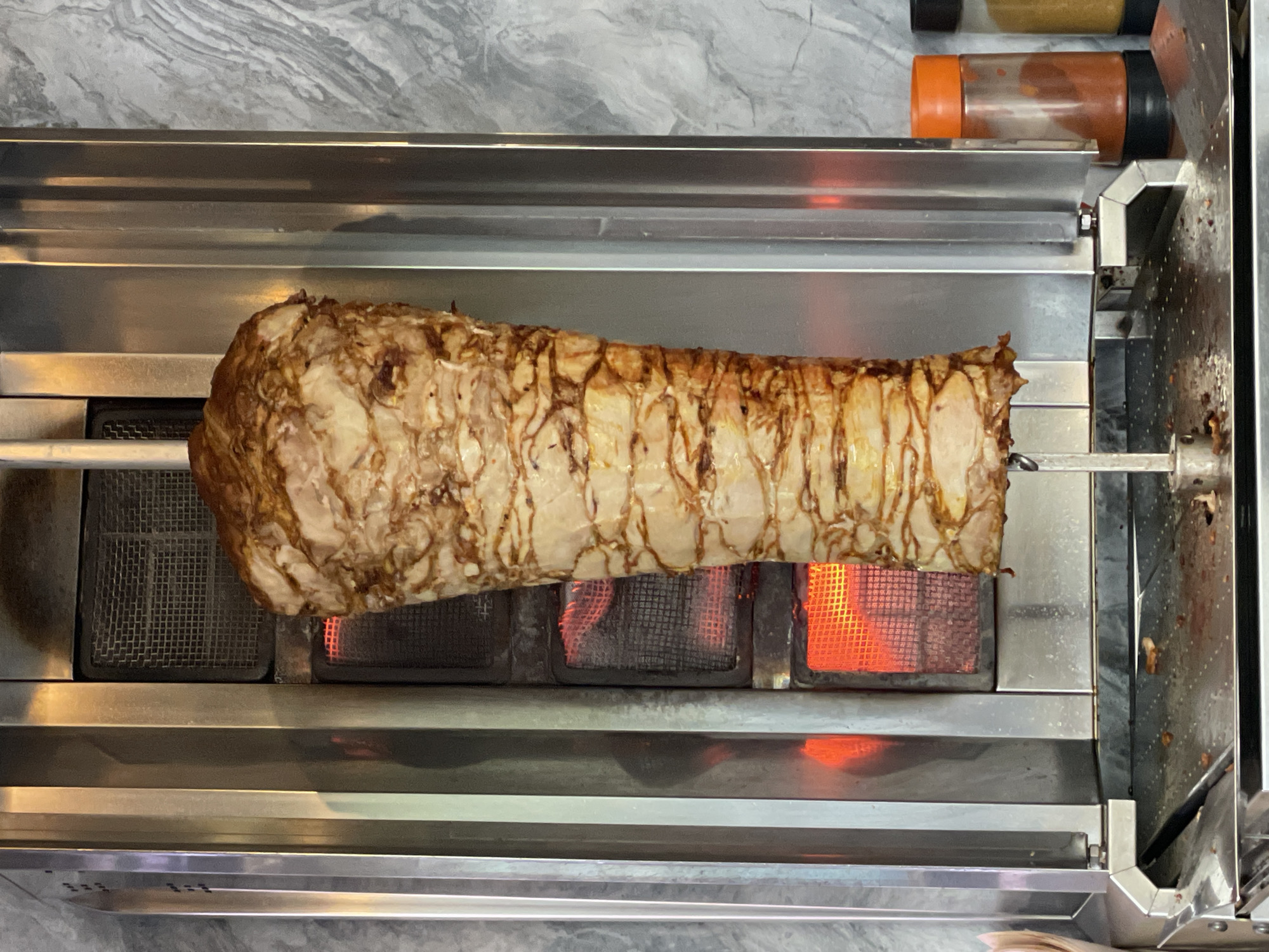 A rotating spit of meat, likely chicken or lamb, is being cooked over a gas flame within a stainless-steel vertical broiler. Condiment containers are visible in the background. The location is not discernible from the image.