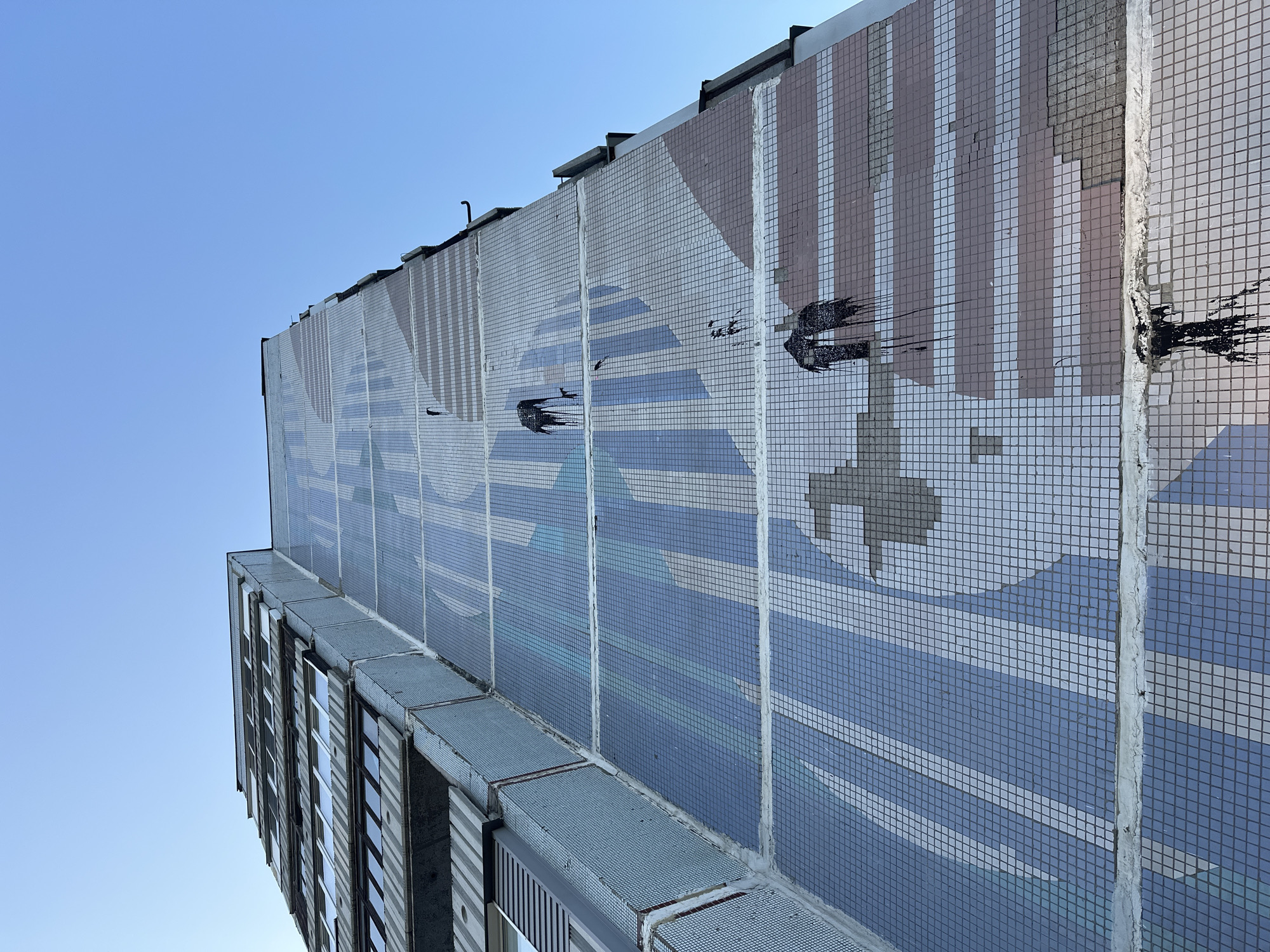 The image is a low-angle shot of the side of a building in Biysk, Russia, featuring a large mosaic mural. The building appears to be a multi-story apartment complex with balconies. The mosaic depicts abstract shapes and stripes in shades of blue, gray, and pink. There are several black, tar-like splatters of paint or some other substance dripping down the mosaic, particularly on the upper right and lower right sections. The sky is clear and blue. The building's facade shows signs of wear and tear, with some mosaic tiles missing or damaged. The overall impression is one of an urban environment with a piece of public art that has been defaced.