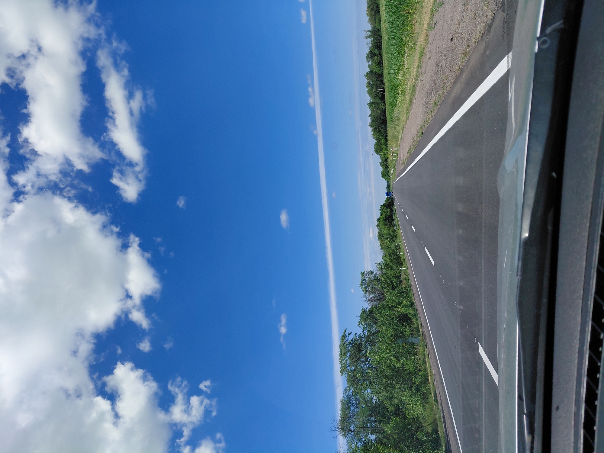 The image shows a view from the perspective of someone inside a vehicle, looking out at a two-lane asphalt road stretching into the distance. The road has clear white lane markings. On the left side of the road, there are dense green trees. On the right side, there is a field of tall green crops, likely corn, and further in the distance, more trees line the horizon. The sky is a bright blue with scattered white cumulus clouds. The lower part of the image shows the dashboard and windshield of the vehicle, with the windshield wipers visible. There are no people or animals visible in the image. The scene appears to be a rural or semi-rural area on a sunny day.