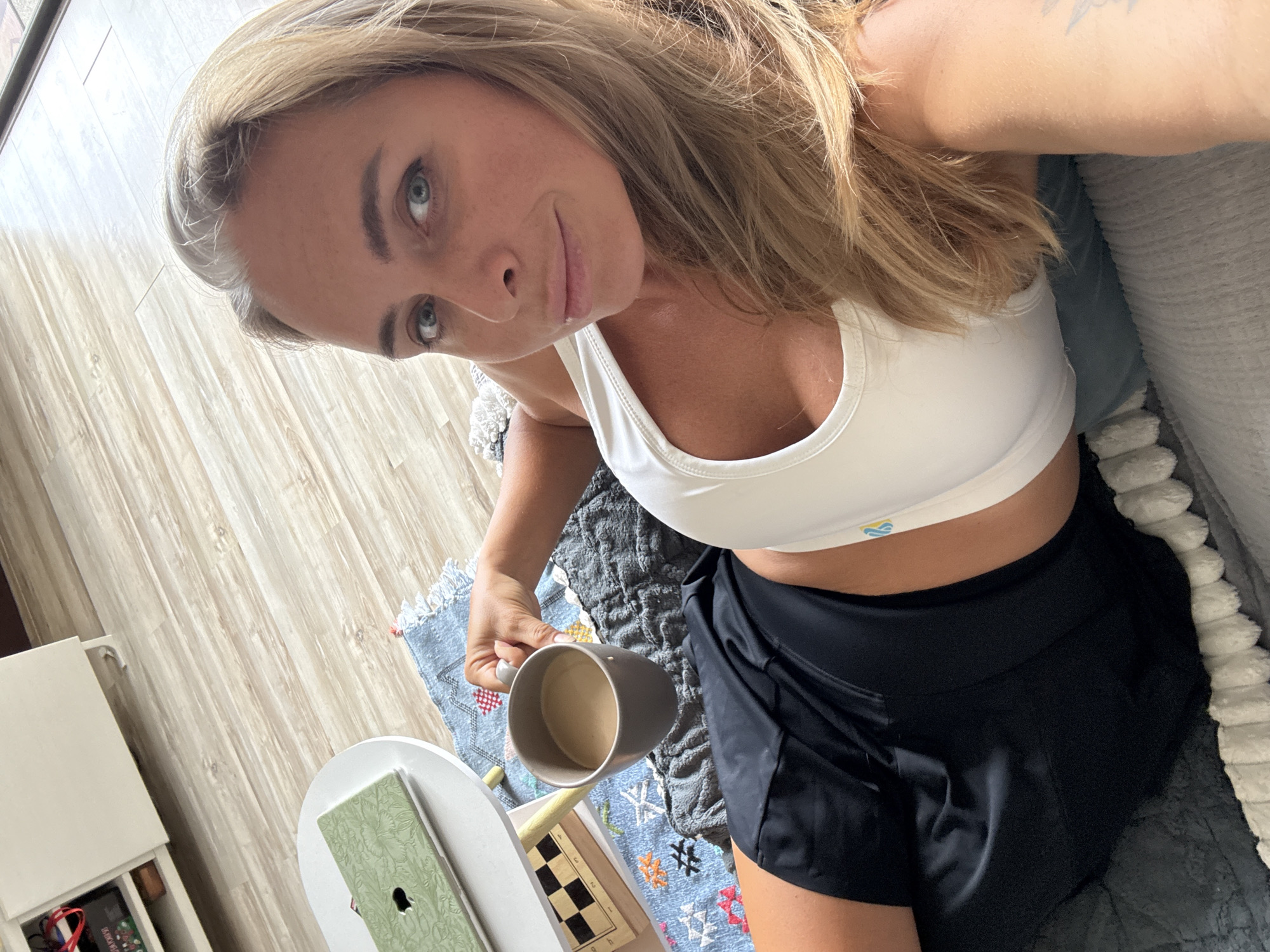 A woman is lying down, holding a mug of coffee. She is wearing a white sports bra and black shorts. The background shows a wooden floor and a white cabinet. There is also a small table with a laptop and a chessboard on it. The scene appears to be an indoor setting, possibly a home, in Ko Samui District, Thailand.