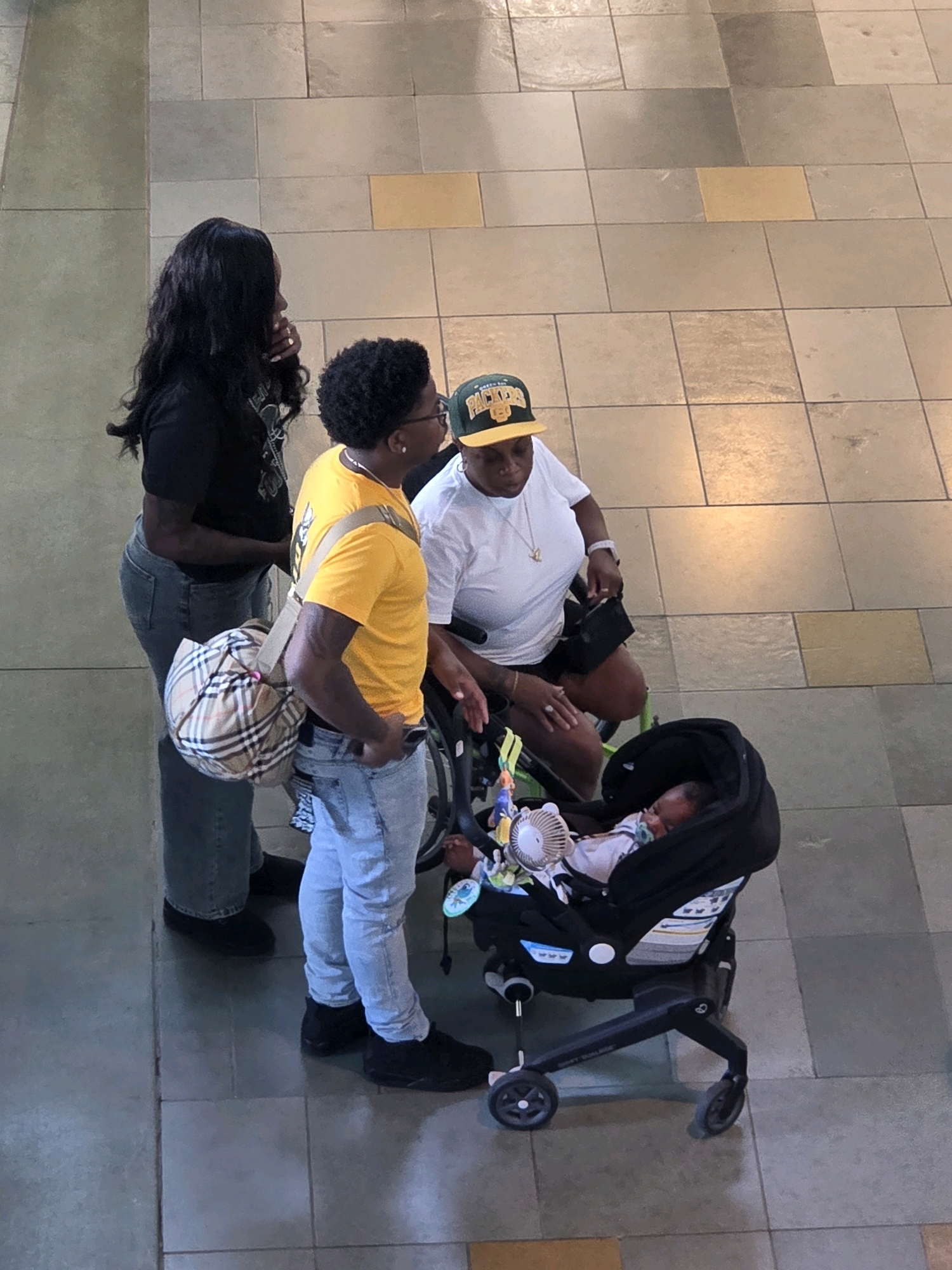 Four individuals are standing on a tiled floor in an urban environment. A woman with long black hair, wearing a black t-shirt and jeans, is on the left. Next to her is a man wearing a yellow t-shirt, light blue jeans, and black sneakers, carrying a plaid bag over his shoulder. To the right of the man, a woman wearing a white t-shirt and black shorts, and a green and yellow baseball cap, is seated in a wheelchair. In front of the seated woman, a baby is sleeping in a black stroller. The tiled floor consists of large square tiles of varying shades of gray and some yellow accents. The background appears to be a neutral-colored wall. The scene suggests a public space, potentially a shopping mall or an outdoor plaza, located in Long Beach, United States of America.