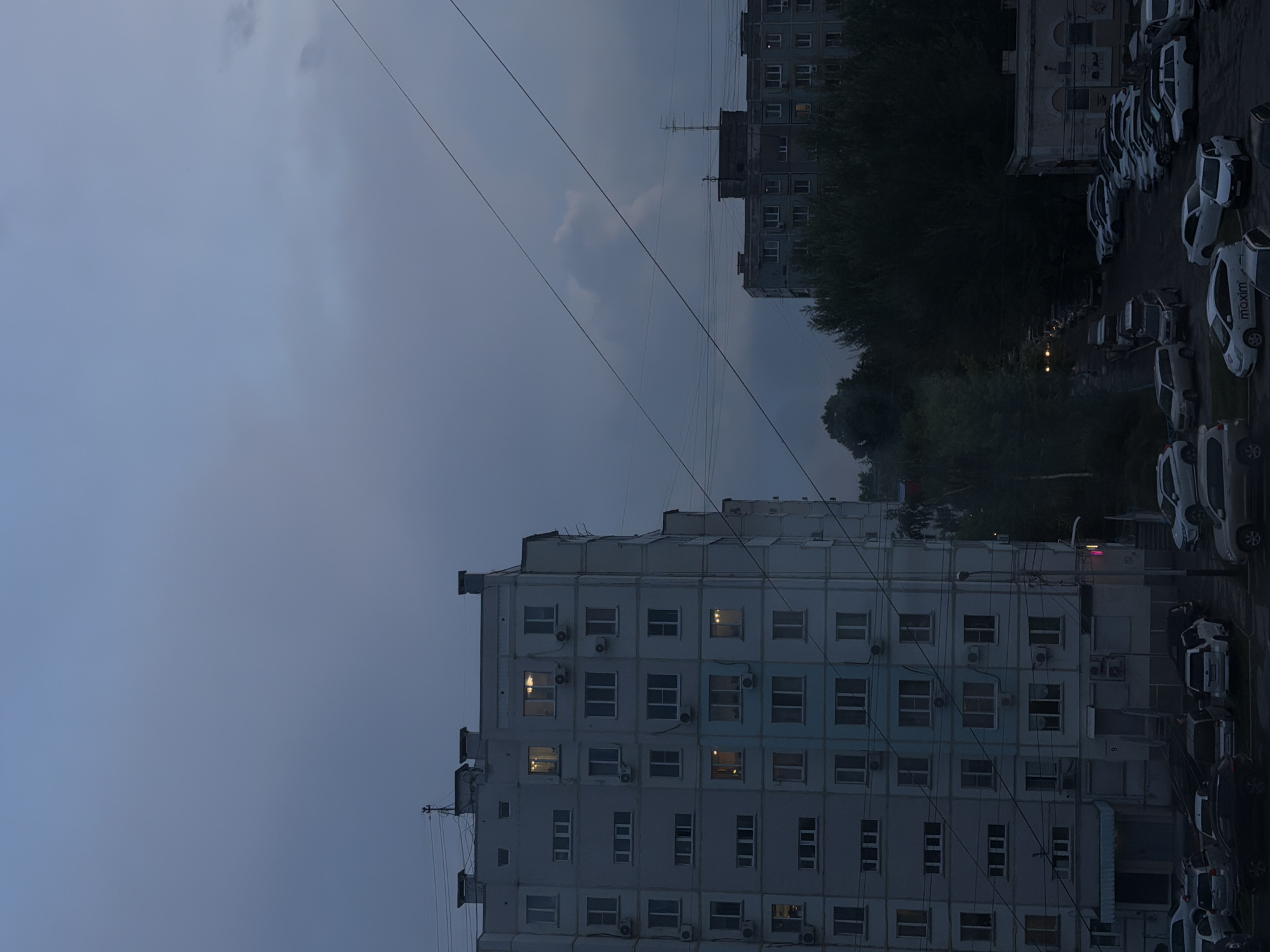 The image depicts a nighttime view of a residential area in Khabarovsk, Russia. The scene is dominated by several multi-story apartment buildings, characteristic of Soviet-era architecture, with windows lit from within. Power lines crisscross the sky, which is a deep, muted blue, suggesting twilight or overcast conditions. In the foreground and mid-ground, a street and parking lot are visible, filled with a variety of parked cars. The overall atmosphere is quiet and urban. The buildings appear to be concrete structures with a light-colored facade, some with visible air conditioning units on their exteriors. Trees and greenery are sparsely scattered between the buildings and along the edges of the scene, adding a touch of nature to the urban landscape. The ground is wet, indicating recent rain or damp conditions. No people are visible in the frame. The lighting is dim, with artificial lights from the buildings and streetlamps casting a soft glow on the surroundings.