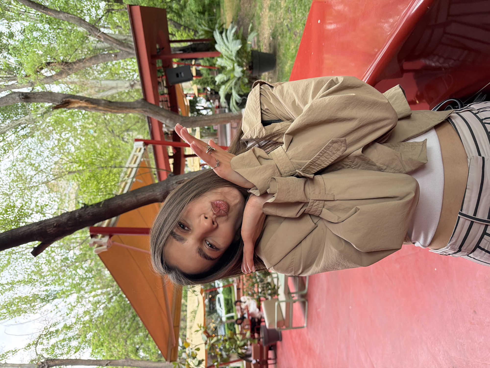 A young woman is pictured in an outdoor setting with lush greenery. She is wearing a tan-colored jacket, a white top, and vertically striped pants. She is making a "kiss" face with her lips pursed. Her hands are raised towards her face, and she is wearing rings on her fingers. The background features trees, plants, and a red surface, possibly a part of the outdoor setting. The location appears to be Yerevan, Armenia.