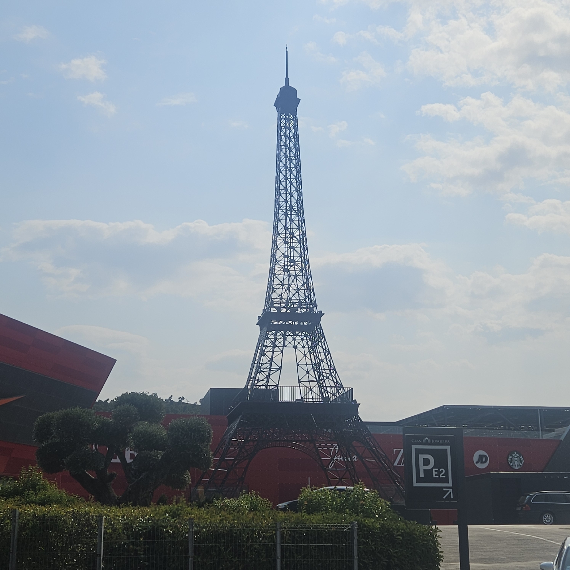 The image depicts a replica of the Eiffel Tower situated in La Jonquera, Spain. The tower is constructed of dark metal lattice and stands tall against a bright, partly cloudy sky. In the foreground, there is lush green foliage and a metal fence. Behind the foliage and to the left of the tower, a modern red building with a geometric design is visible. To the right of the tower, a large building with a dark, slanted roof is present, featuring signage that includes "GRAN JONQUERA", a parking symbol indicating "P E2", the JD logo, and the Starbucks logo. A black car is parked to the right of this building. The overall scene suggests an outdoor commercial or entertainment area.