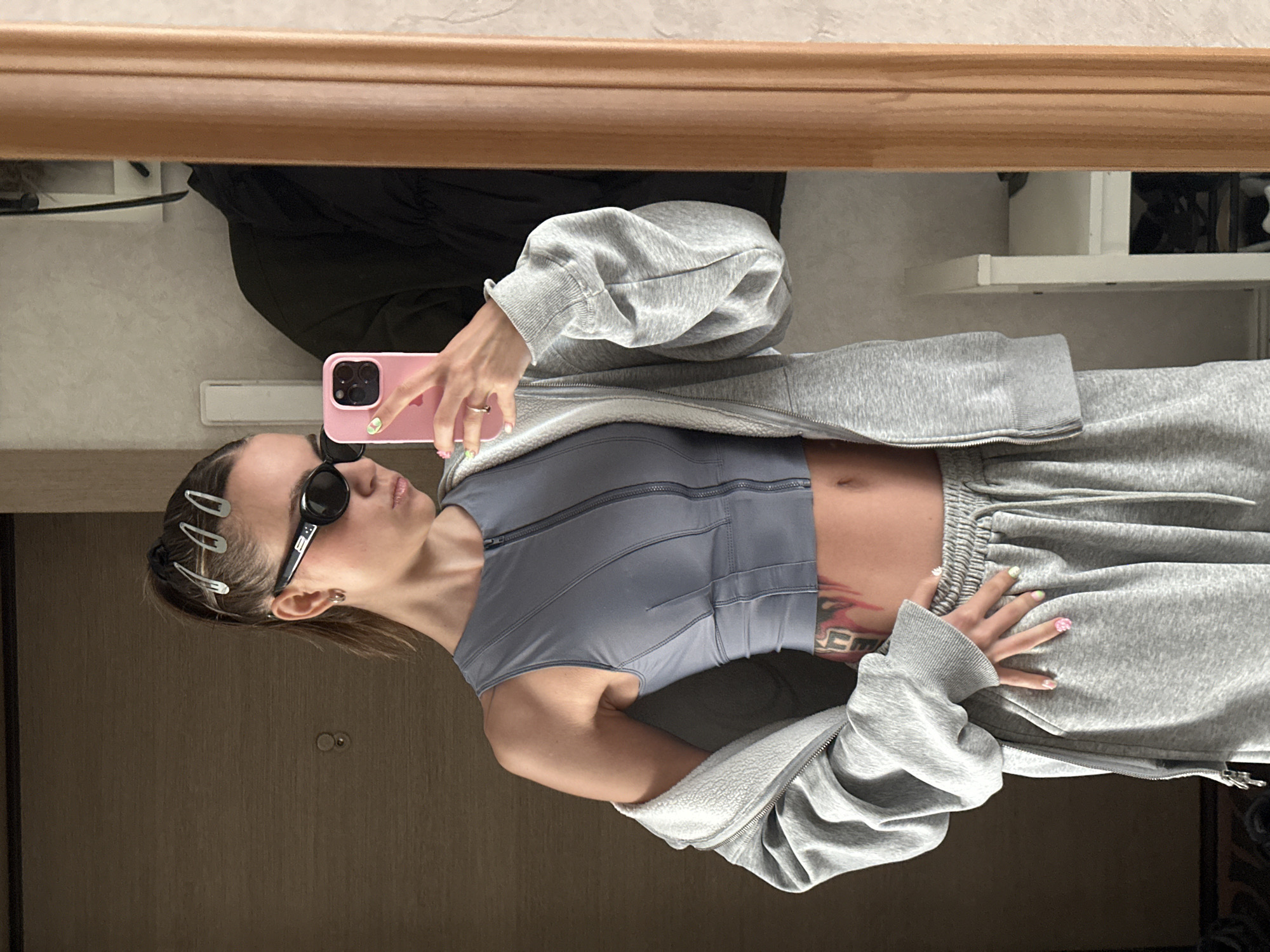 In this vertical, slightly blurry selfie taken in Moscow, Russia, a young woman is captured in a relaxed pose. She is wearing a gray zip-up hoodie, a grey sports bra, and grey sweatpants. The woman has dark hair pulled back, with several silver hair clips adorning her head. She is also wearing black sunglasses and a silver hoop earring in her visible ear.
She is holding a pink smartphone in her right hand, which is reflecting in a mirror that frames the top of the image. Her left hand is resting on her sweatpants, revealing colorful nail art with floral designs. A large tattoo is visible on her abdomen.
The background, seen in the mirror's reflection, suggests an indoor setting, possibly a room in an apartment or hotel, with hints of furniture, shelves, and clothing. The lighting appears to be ambient indoor light, with no strong indications of time of day or weather conditions. The overall mood is casual and laid-back. No visible text is present in the image.